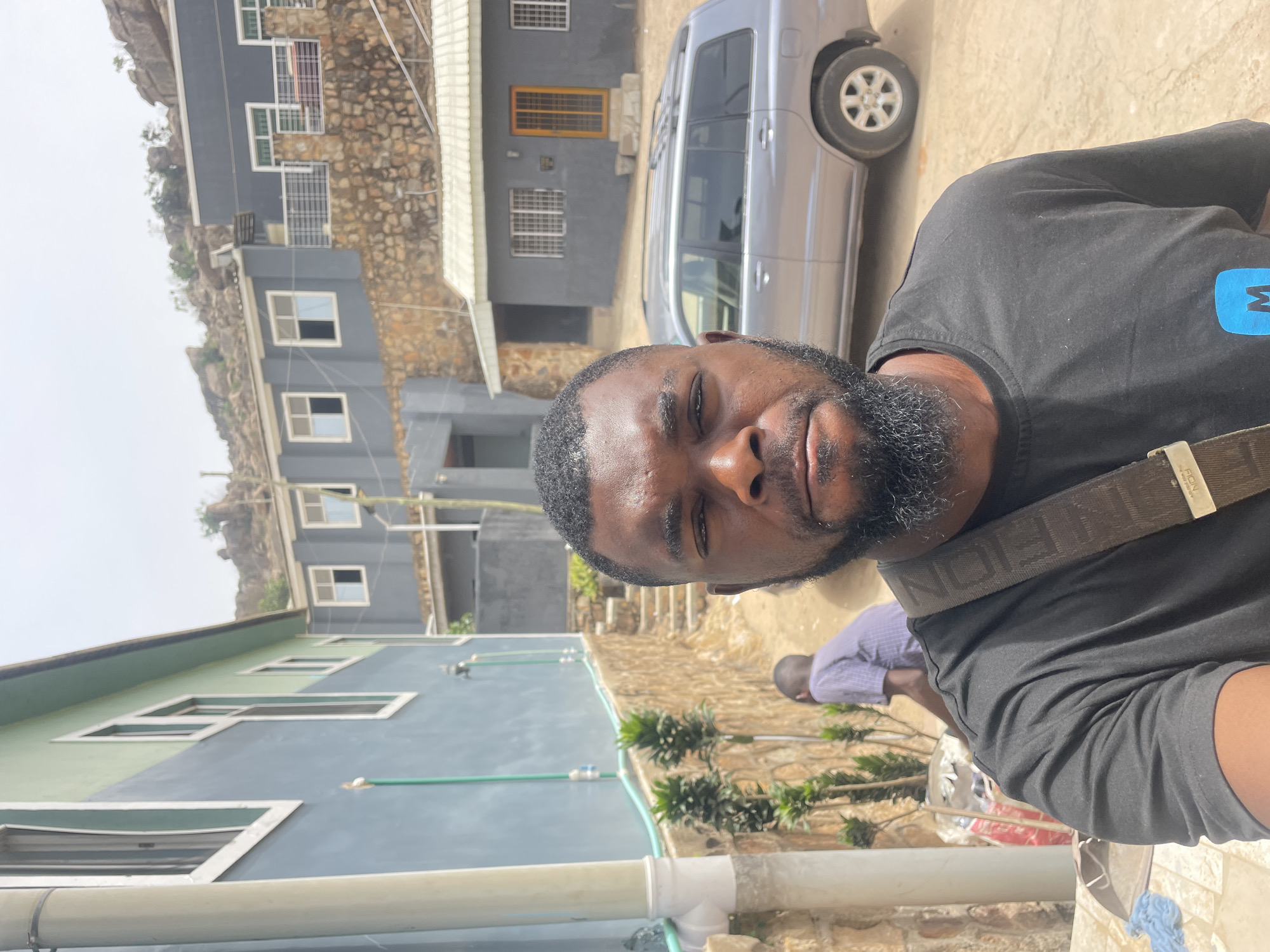 A man with a dark beard and mustache is prominently featured in a selfie, looking directly into the camera. He wears a black short-sleeved t-shirt with a blue logo on the right side of his chest and a brown shoulder strap bag, which appears to have "FENDI" written on it.
The background reveals a residential scene in Bukuru, Nigeria, under bright daylight with clear skies. Several multi-story buildings, predominantly grey and light green/blue in color, are visible. These structures are built into or alongside rocky, hilly terrain. The buildings feature white-framed windows, some with security bars, and exterior piping for utilities. A silver SUV is parked on a dirt path behind the man and to his right. Further back, another individual, seen from behind in a light-colored shirt, appears to be engaged in an activity on the ground. Small green plants and bushes are scattered throughout the scene. The ground is a mix of paved and unpaved dirt. The man's expression is neutral.