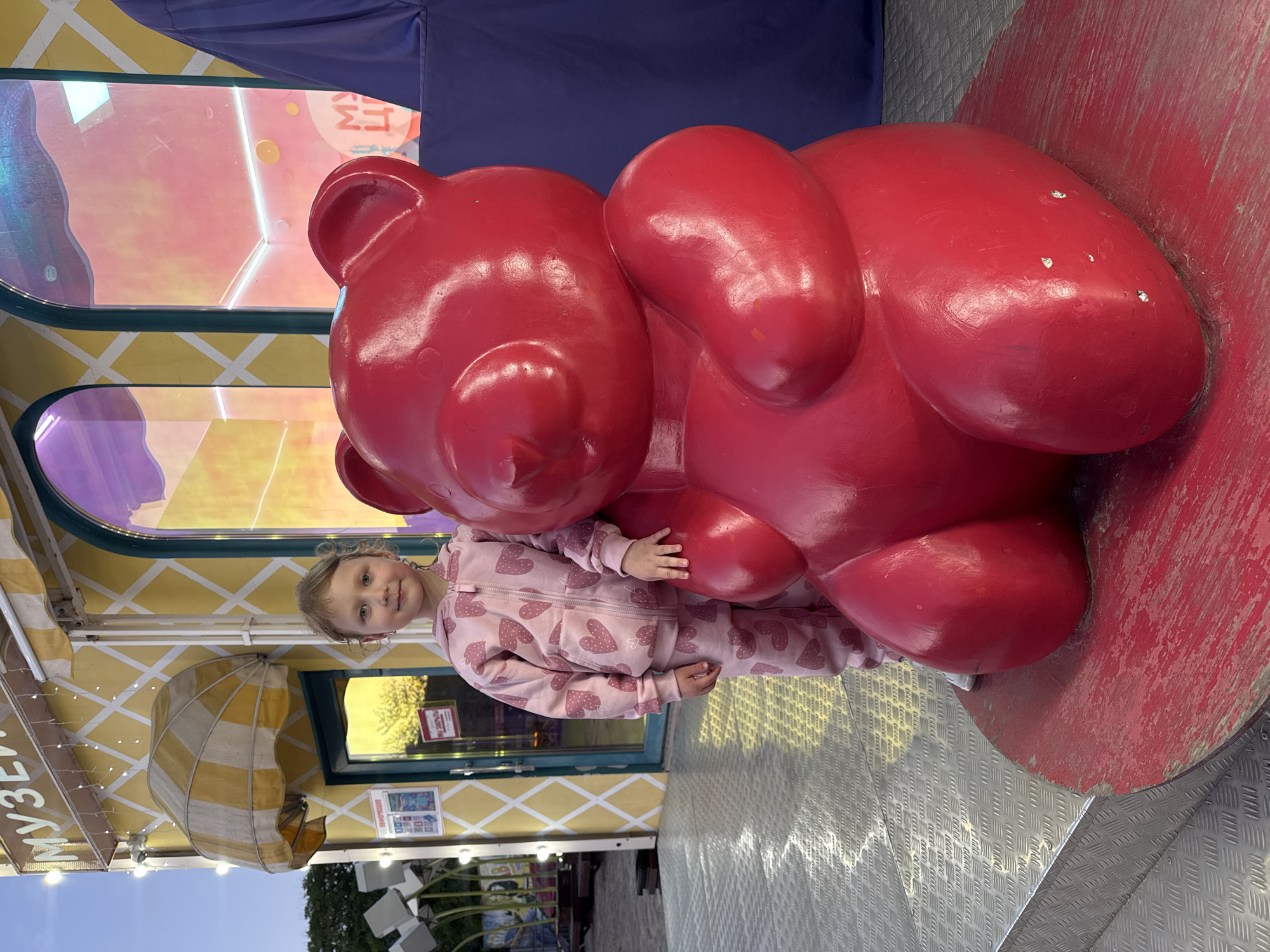 A young girl in a pink heart-patterned tracksuit stands beside a large, red, gummy-bear-shaped sculpture. The sculpture rests on a circular red platform. The scene is outdoors, with a yellow building with white stripes and patterned windows in the background. A sign partially visible reads "MY3E". The foreground features a metallic, patterned surface, and the sky is visible in the lower left. The location is Yevpatoriya, Ukraine.
