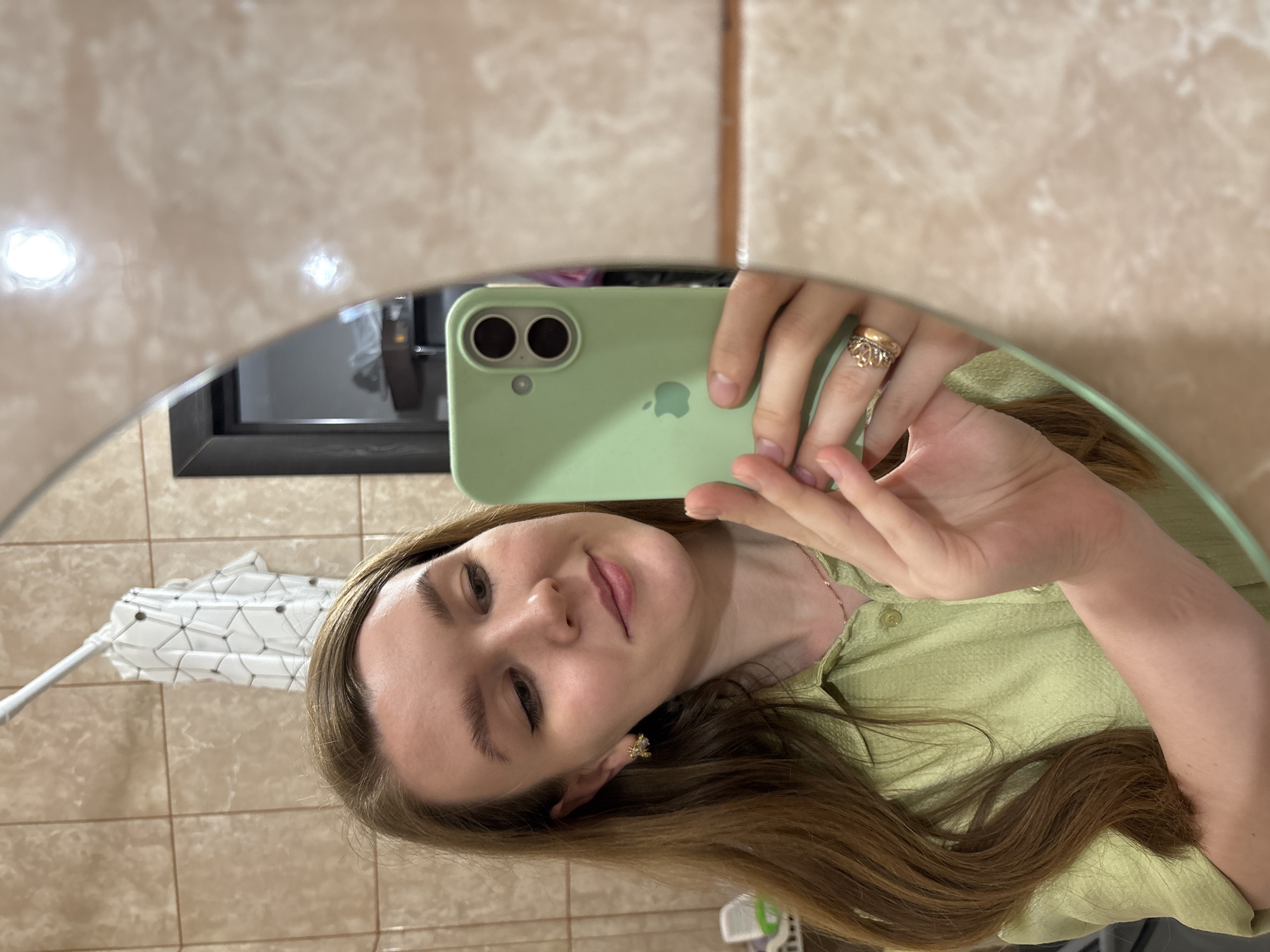 A selfie is taken in a bathroom. A young fair-skinned woman with long brown hair is reflected in a round mirror. She is holding a green phone with a light green phone case. Her right hand is raised, making a peace sign. She is wearing a light green button-down shirt and a gold ring. Behind her, the bathroom features beige tiling, a black-framed window, and a white duster. It is not possible to determine the location in Izhevsk, Russia based on the provided image.
