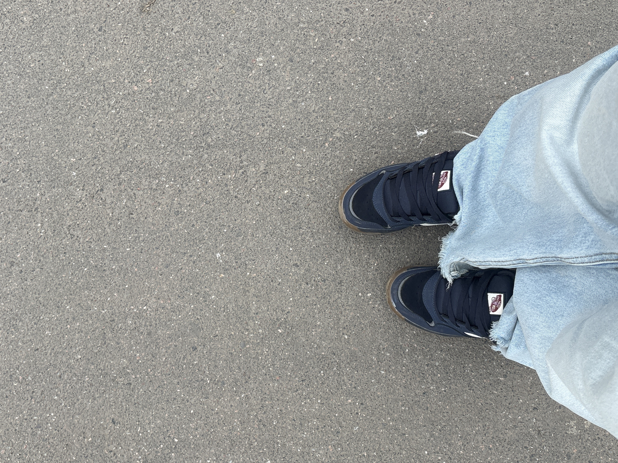 An overhead shot captures a pair of feet wearing dark blue sneakers and light blue, distressed denim jeans, standing on a textured asphalt surface. The feet are positioned in the lower right quadrant of the frame, with ample grey asphalt occupying the rest of the space.
The sneakers feature dark blue uppers, thick white laces, and distinct brown rubber soles. On the tongue of both shoes, a white rectangular label with red trim is visible, bearing the "OFF THE WALL" text above the "VANS" logo. The light blue jeans have a casual, worn look, with frayed edges around the ankles, partially covering the top of the shoes.
The asphalt ground is composed of small, varied grey and black pebbles, creating a rough, granular texture, typical of a sidewalk, street, or parking lot. The lighting appears even, suggesting an overcast day or indirect light, with no prominent shadows. There is no discernible activity beyond standing. While the provided context states Saint Petersburg, Russia, there are no visual elements within the image itself to confirm this specific location.