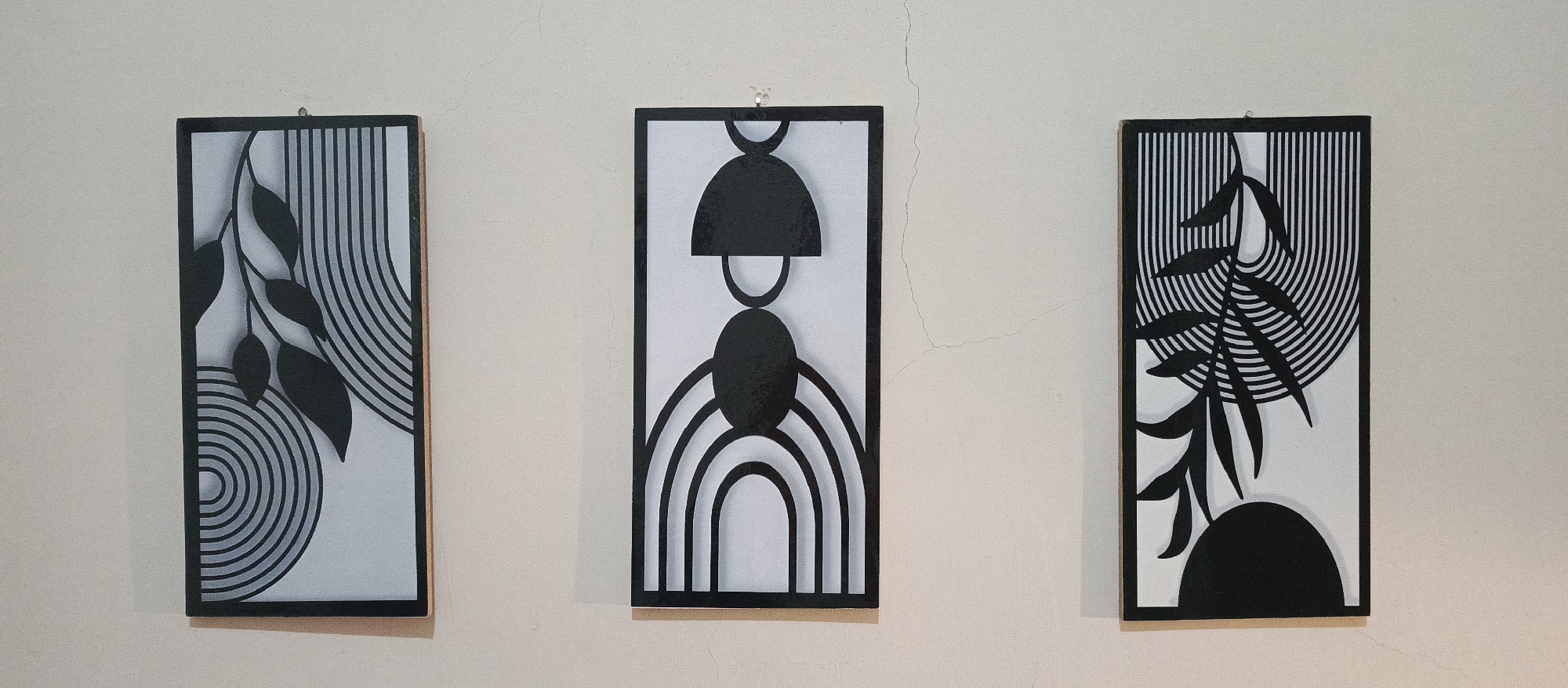 Three rectangular artworks with black and white abstract designs are displayed on a light-colored wall. The artwork on the left features a branch with leaves and concentric arcs. The central artwork depicts a symmetrical arrangement of shapes, including a semicircular top, a circular middle, and rainbow-like arcs at the bottom. The artwork on the right shows a branch with leaves against a background of vertical lines and concentric arcs. The wall has visible cracks.
