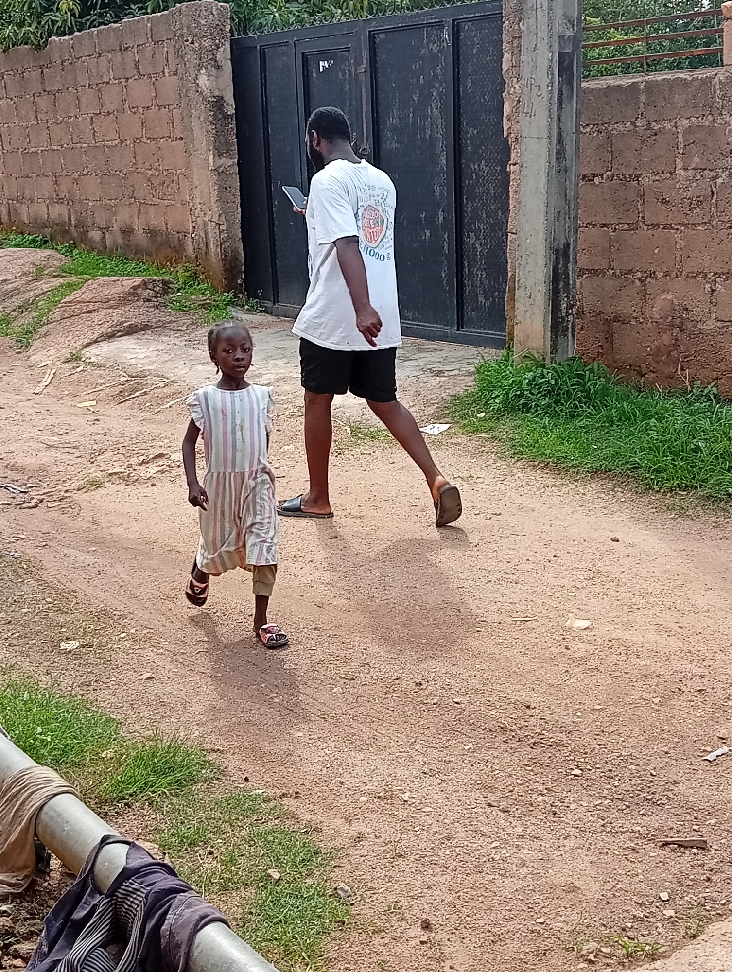 A young girl in a striped dress and sandals walks on a dirt path in what appears to be an outdoor urban environment. Behind her, a man in a white t-shirt and black shorts, holding a phone, walks away from the camera. The background features a brick wall, a large black metal gate, and lush green vegetation. The ground is composed of reddish-brown dirt with scattered stones and a few pieces of debris. A metal pipe with some clothing draped over it is visible in the lower left foreground. The lighting suggests it is daytime. The scene is likely located in Jos, Nigeria, given the visual cues of the architecture and environment.