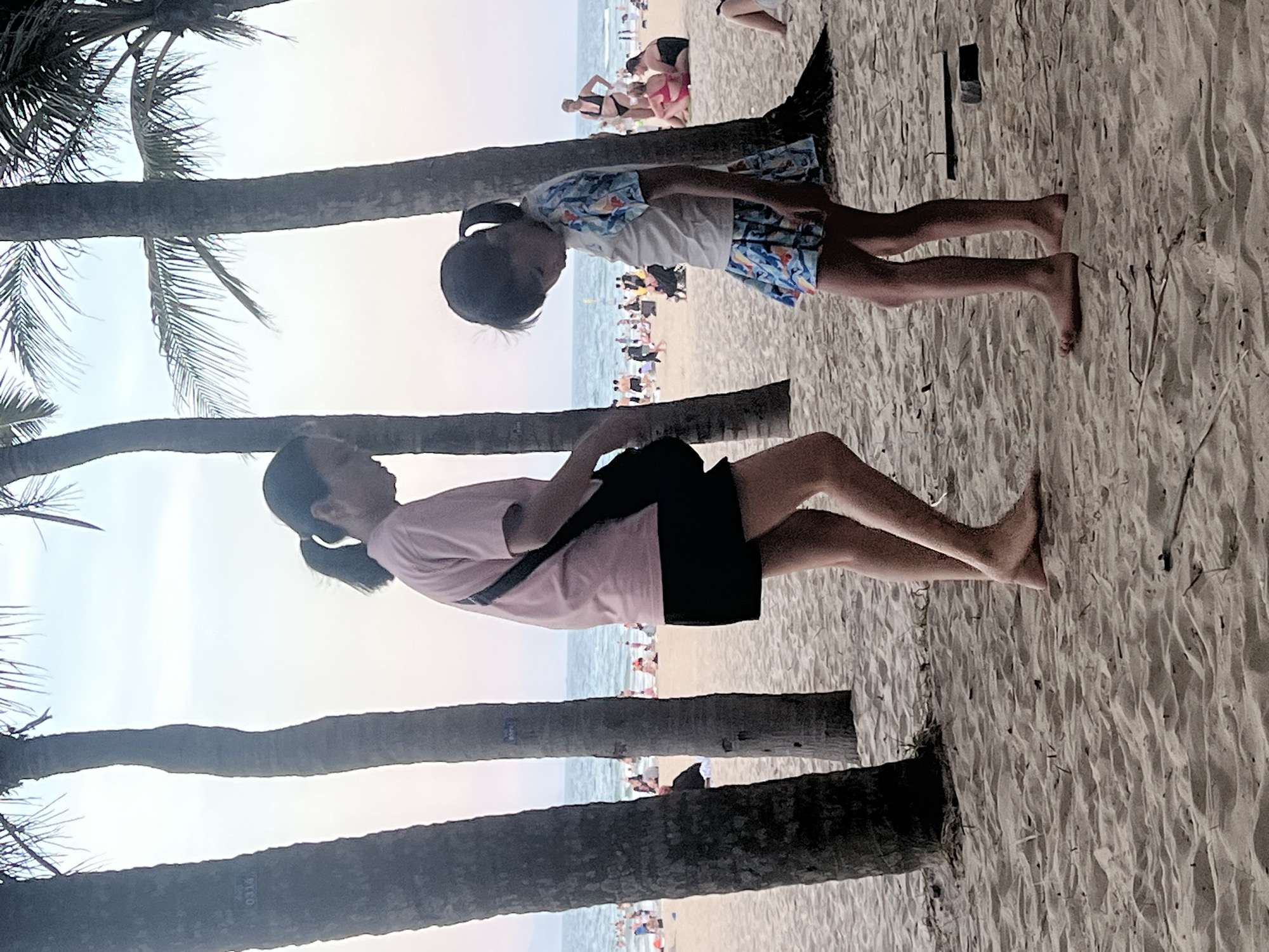 The image depicts a beach scene in Da Nang, Vietnam, likely during the late afternoon or early evening, given the soft lighting. In the foreground, two individuals are present. On the left, an adult, appearing to be female, is wearing a pink t-shirt and a black skirt. She has dark hair pulled back in a ponytail and is carrying a dark bag over her shoulder. She is looking down. To her right, a child, also appearing to be female, is wearing a white t-shirt with a colorful pattern and a matching patterned skirt. She has dark hair styled in a ponytail and is looking downwards. Both are standing on a sandy beach with palm trees visible behind them, their trunks dark and textured. The background reveals a crowded beach with numerous people in swimwear, some sitting and others walking. The ocean is visible, with gentle waves and the horizon line under a pale, hazy sky. The sand is light brown and appears well-trodden, with some small debris scattered on it. The overall atmosphere suggests a relaxed, public recreational setting.