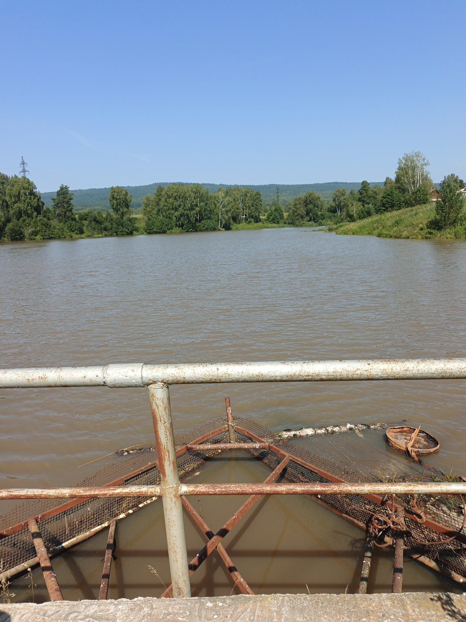 The image shows a view from a concrete embankment overlooking a wide, muddy-brown river. In the foreground, there is a rusty metal railing and a section of a concrete structure. Partially submerged in the water and attached to the embankment are various pieces of rusted metal structures and netting, possibly part of a fishing weir or some form of water management system. Some debris, including what appears to be a circular metal object and some plant material, is visible in the water around these structures.
The river extends into the distance, bordered by a lush green shoreline with a dense forest of deciduous trees and some evergreen trees. Beyond the immediate treeline, rolling hills covered in dark green vegetation are visible under a clear, bright blue sky. A power line pylon is faintly visible in the distance on the left side of the frame. The overall scene depicts a natural landscape with signs of human infrastructure integrated into it. The location is stated to be the city of Urtiushka, Russia.