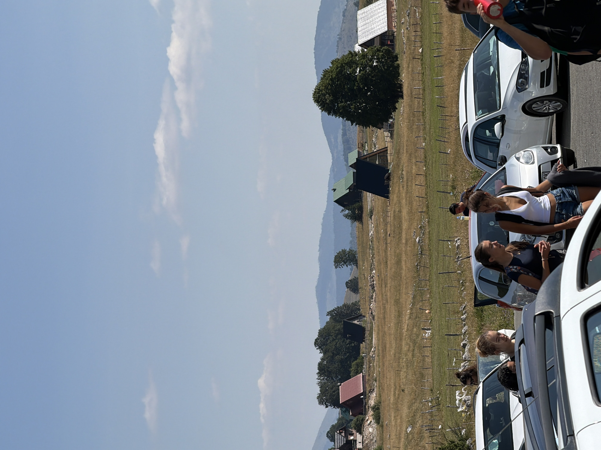 The image captures a sunny daytime scene in Virak, Montenegro, with a focus on a parking area adjacent to a picturesque landscape. In the foreground, several white and silver cars are parked, partially obscuring a group of people. One woman, wearing a white tank top and denim shorts, is standing near a white car. To her right, another woman in a dark shirt with a backpack is visible. Further back, a white station wagon is parked, with people in and around it.
The background reveals a vast, dry, golden field with sparse vegetation and a wooden fence. Several A-frame style houses with dark roofs and some conventional houses with red and white roofs are scattered across the field and on rolling hills. Mature green trees dot the landscape, and further in the distance, several layers of blue-toned mountains recede into the horizon. The sky is a clear, pale blue with a few wispy white clouds. The overall impression is one of a rural setting, likely a popular tourist or recreational area given the presence of multiple vehicles and people.