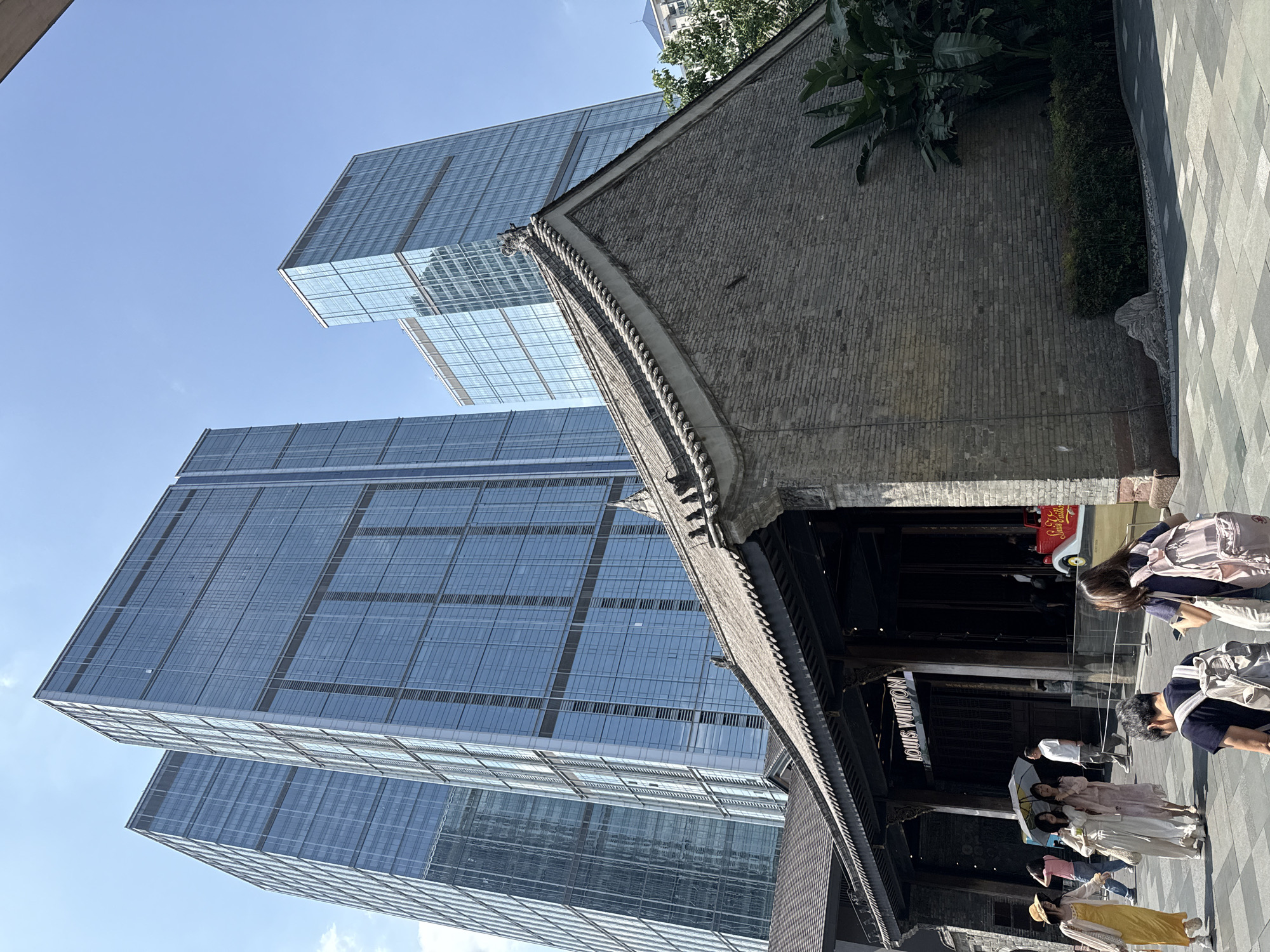 The image captures a street scene in Jinjiang District, China, showcasing a juxtaposition of architectural styles. In the background, modern skyscrapers with glass facades rise against a clear blue sky. In the foreground, a traditional Chinese building with a dark-tiled roof and intricate wooden detailing stands. People, dressed in a variety of modern clothing, walk along the paved street. The entrance to a "LOUIS VUITTON" store is visible, blending the traditional facade with a modern luxury brand. The overall impression is a blend of old and new, indicative of Jinjiang District's urban landscape.