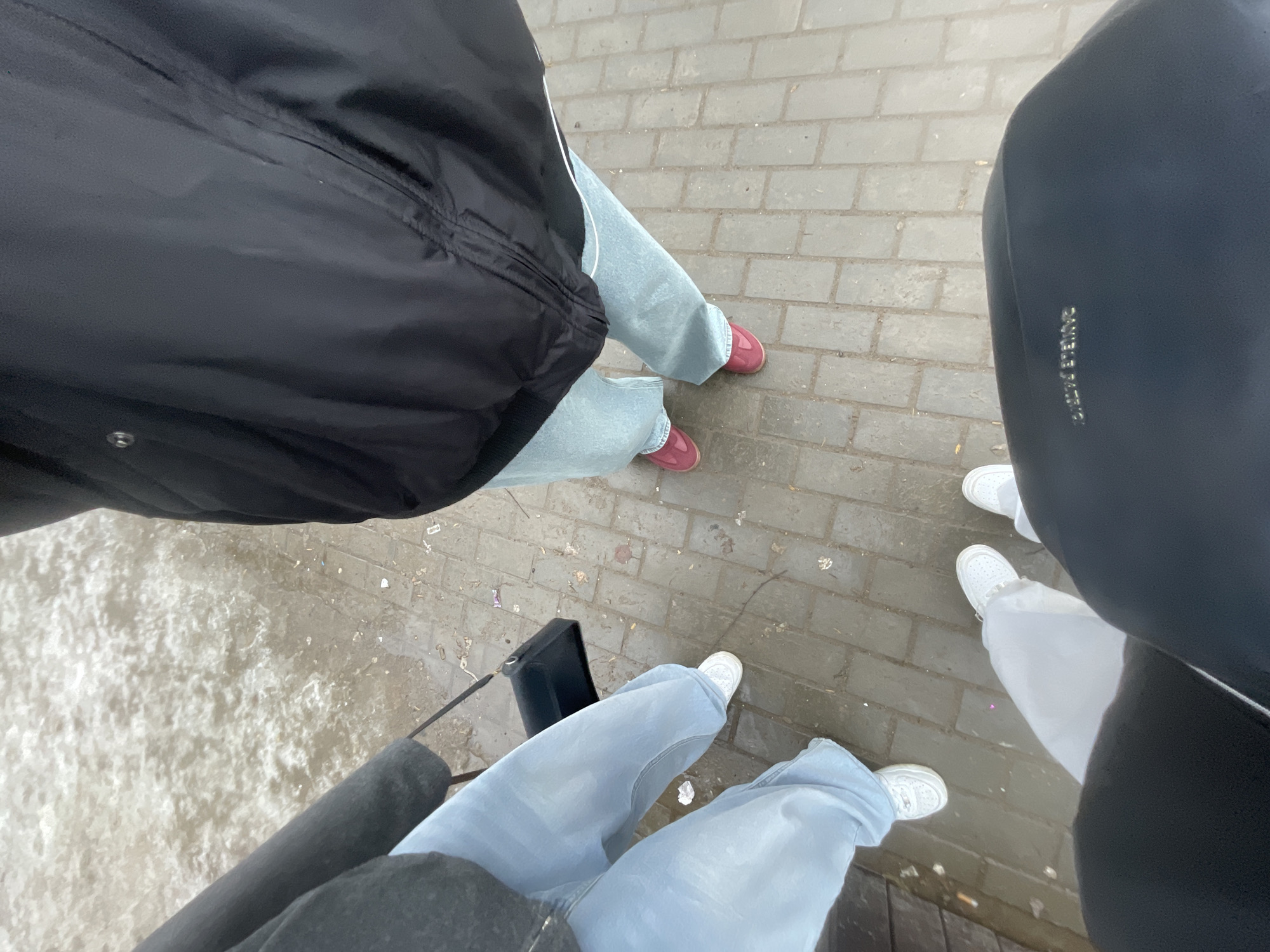 A high-angle photograph depicts the lower bodies of three individuals standing on a wet, light gray paver-brick sidewalk in Chaikovsky, Russia. To the far left, an adjacent area is covered with dirty, partially melted snow and ice.
Subject One, positioned on the right side of the frame, wears a black jacket, light blue wide-leg jeans, and glossy dark red flat shoes.
Subject Two, located on the left side of the frame, wears a dark gray coat, light blue wide-leg jeans, and white sneakers. A black shoulder bag with a thin strap hangs from their left hand at thigh level.
Subject Three, likely the photographer, is visible from a low vantage point in the foreground. This individual wears white pants and white sneakers. A large black bag, potentially a backpack or tote, with partially legible gold lettering "CHANEL PATTON" is visible in the bottom right corner, belonging to Subject Three.
The paved surface is visibly wet, showing puddles and reflections. Small fragments of white litter, possibly paper or plastic, are scattered on the ground. The presence of melted snow and wet paving indicates a damp, likely cold or thawing outdoor environment.