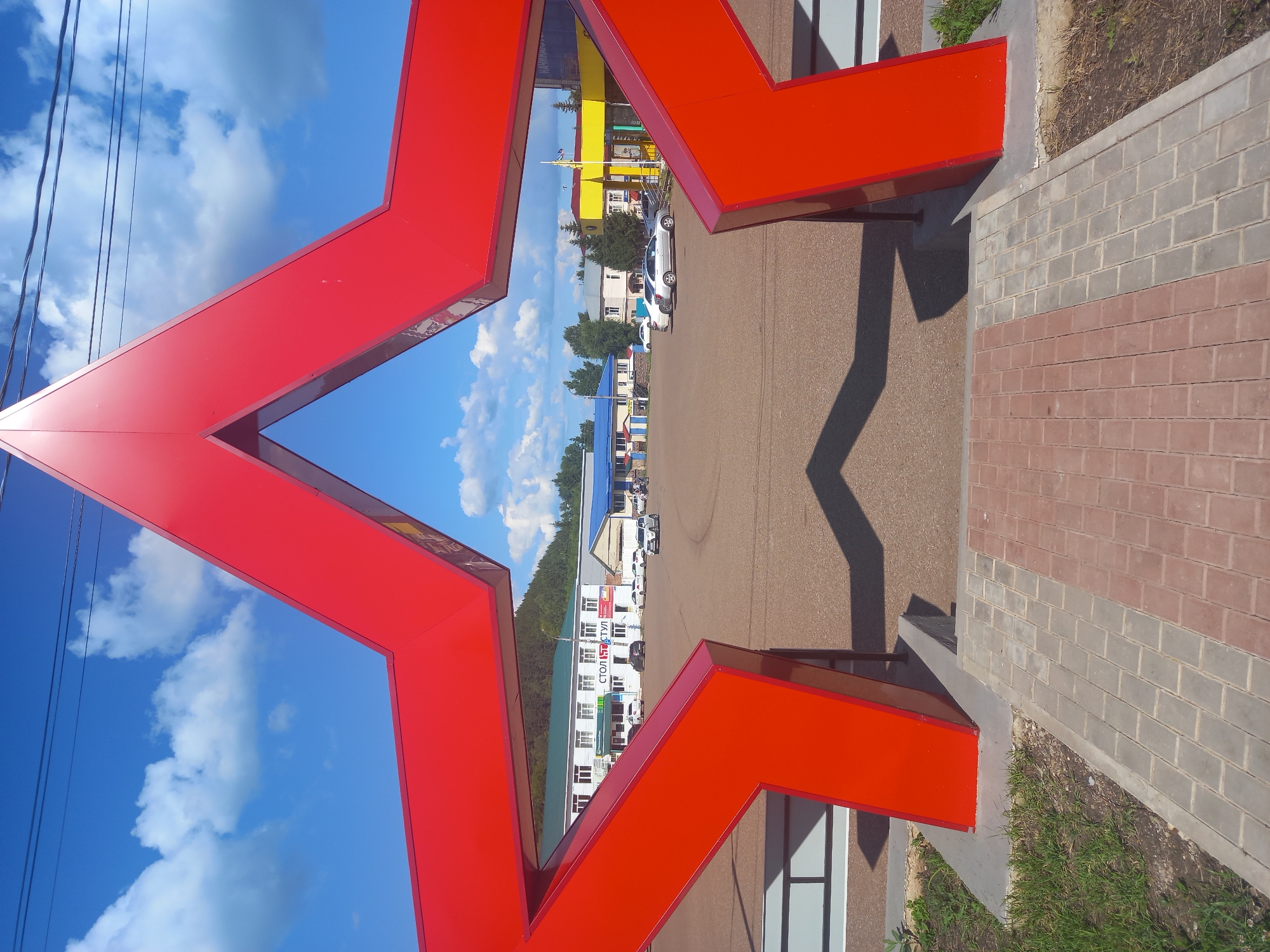 The image shows a large, bright red five-pointed star sculpture framed in the foreground. The star is viewed from an angle that makes it appear hollow, with the sky and a town visible through its opening. The background features a gravel-covered area, several buildings with different colored roofs (white, green, blue, and yellow), parked cars, and a distant treeline with a hill. The sky is a vibrant blue with fluffy white clouds. The ground in the immediate foreground is paved with interlocking bricks in a pattern of red and gray. There are no people visible in the image. The location is identified as the town square in Sterlibashevo, Russia, indicated by the text "СТОЛ И СТУЛ" (Table and Chair) visible on one of the buildings, a common business name in Russia.