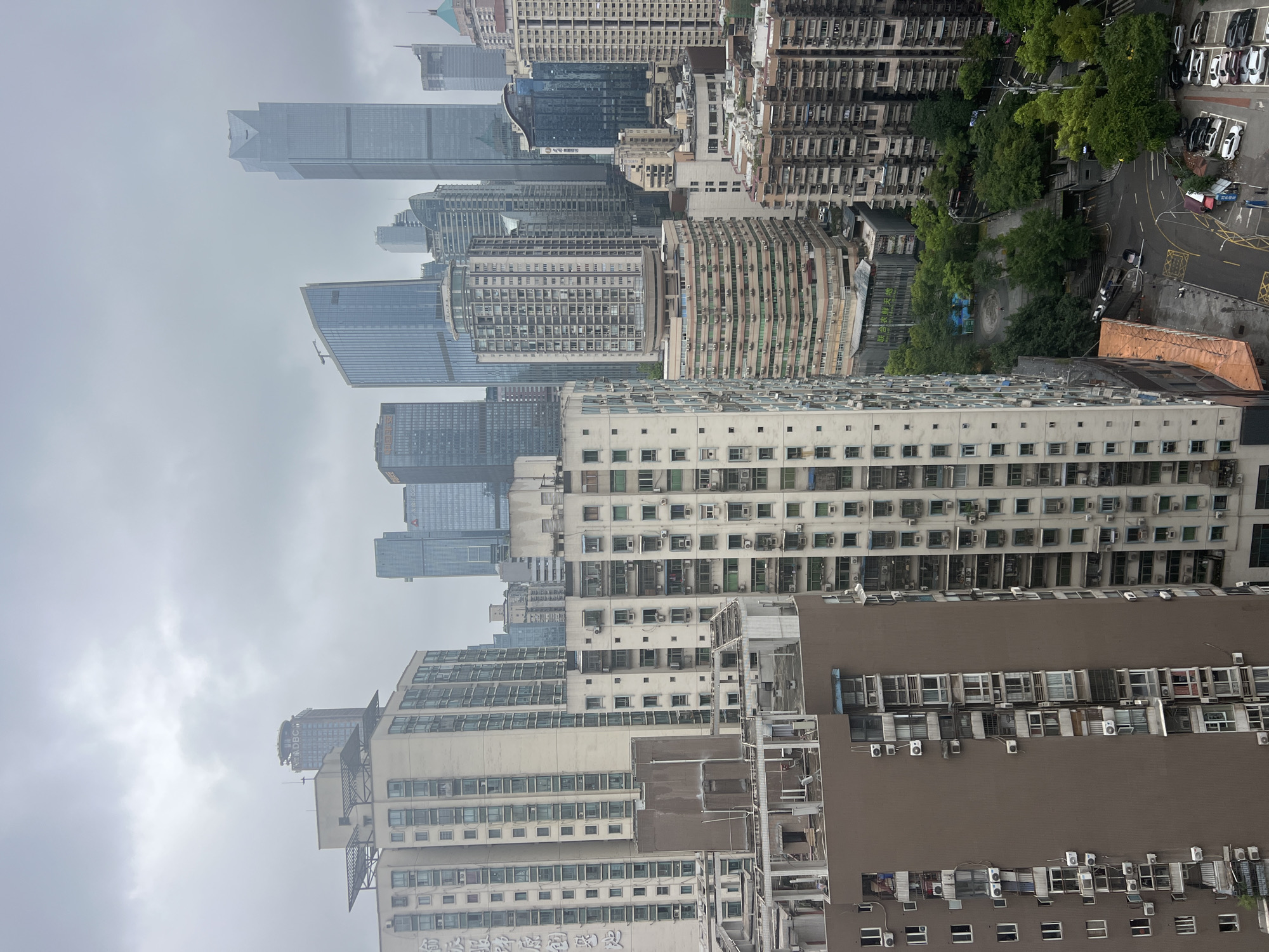 The image shows a dense urban landscape in Yuzhong District, China, characterized by a multitude of high-rise buildings under a cloudy sky. The architecture varies, with some buildings appearing to be residential apartment complexes featuring numerous balconies and air conditioning units, while others are modern skyscrapers with glass facades. The foreground shows a brown residential building with visible windows and air conditioning units. To its right, a lighter-colored residential building with many windows and balconies is visible. In the background, a cluster of taller skyscrapers dominates the skyline, some with distinctive architectural features. A street with parked cars and some greenery is visible at the bottom right, suggesting a bustling city environment. The overall impression is one of a highly populated and developed urban area. No people are clearly discernible in the image.
