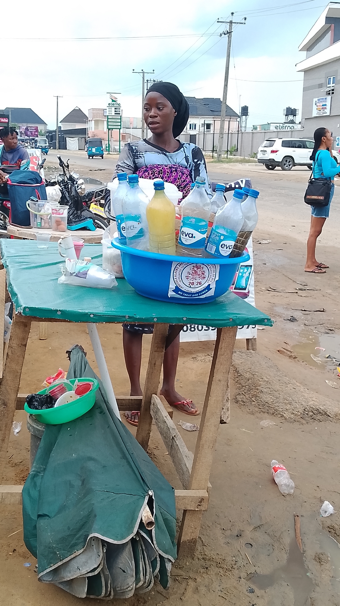 The image depicts a scene in Warri, Nigeria, centered on a female street vendor. The vendor, wearing a head covering and a patterned top, stands behind a makeshift wooden table covered with a green tarp. On the table is a blue plastic basin filled with bottles of water and juice. Next to the basin, there are cups and plastic bags. Underneath the table, there is a folded-up green umbrella and a small green bowl containing dark objects.
The ground is muddy and littered with trash, including discarded plastic bottles. In the background, there are other individuals, motorcycles, and vehicles, including a white SUV. A sign for "eterna" is visible in the distance. A woman in a blue shirt and jean shorts stands in the background.