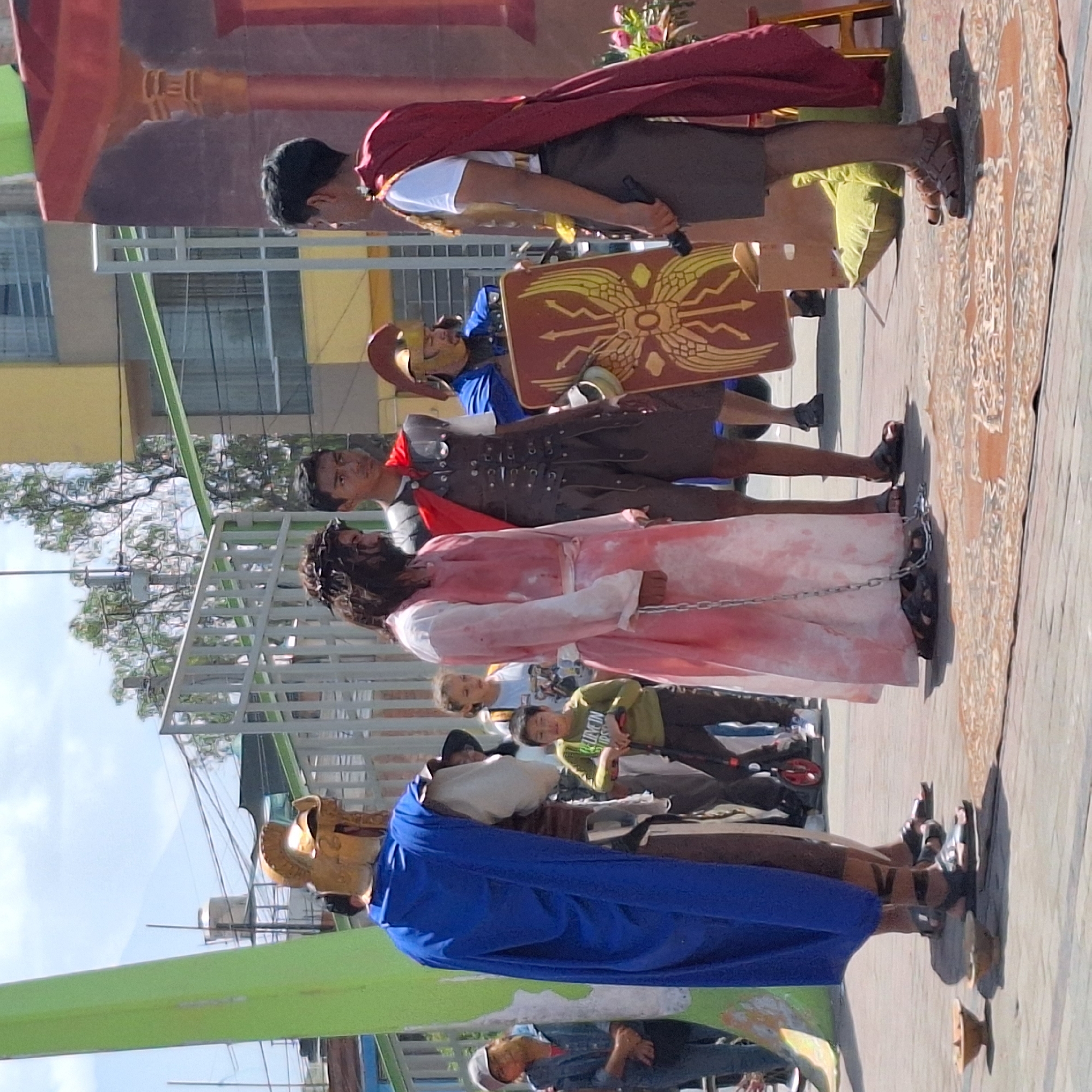 The image depicts a procession of individuals dressed in costumes suggestive of a religious reenactment, likely related to the Passion of Christ. The scene appears to be set in a city location, based on the urban background with buildings and infrastructure.
People and Subjects:
- Several individuals are dressed in costumes that suggest Roman soldiers, complete with helmets, shields, and armor.
- One person is dressed in a white garment with blood-like stains, bound by chains, possibly representing Jesus Christ.
- The figures are walking on a pavement with a decorative pattern.
- Other individuals, possibly onlookers, are present in the background.
Objects:
- Roman soldier costumes, including helmets, shields, and tunics.
- Chains binding the individual in white.
- A decorative patterned pavement.
- A green painted structure with a white fence on the side.
Actions and Interactions:
- The individuals are in procession, walking along the pavement.
- The individuals in costume appear to be portraying characters from a historical or religious event.
- Onlookers appear to be observing the procession.
Environment:
- An urban setting with buildings and streets.
- The presence of streetlights and power lines suggests a populated area.
- The setting could be the city of Mariano Melgar, Peru, given the prompt's instructions, although this is not visually verifiable in the image.