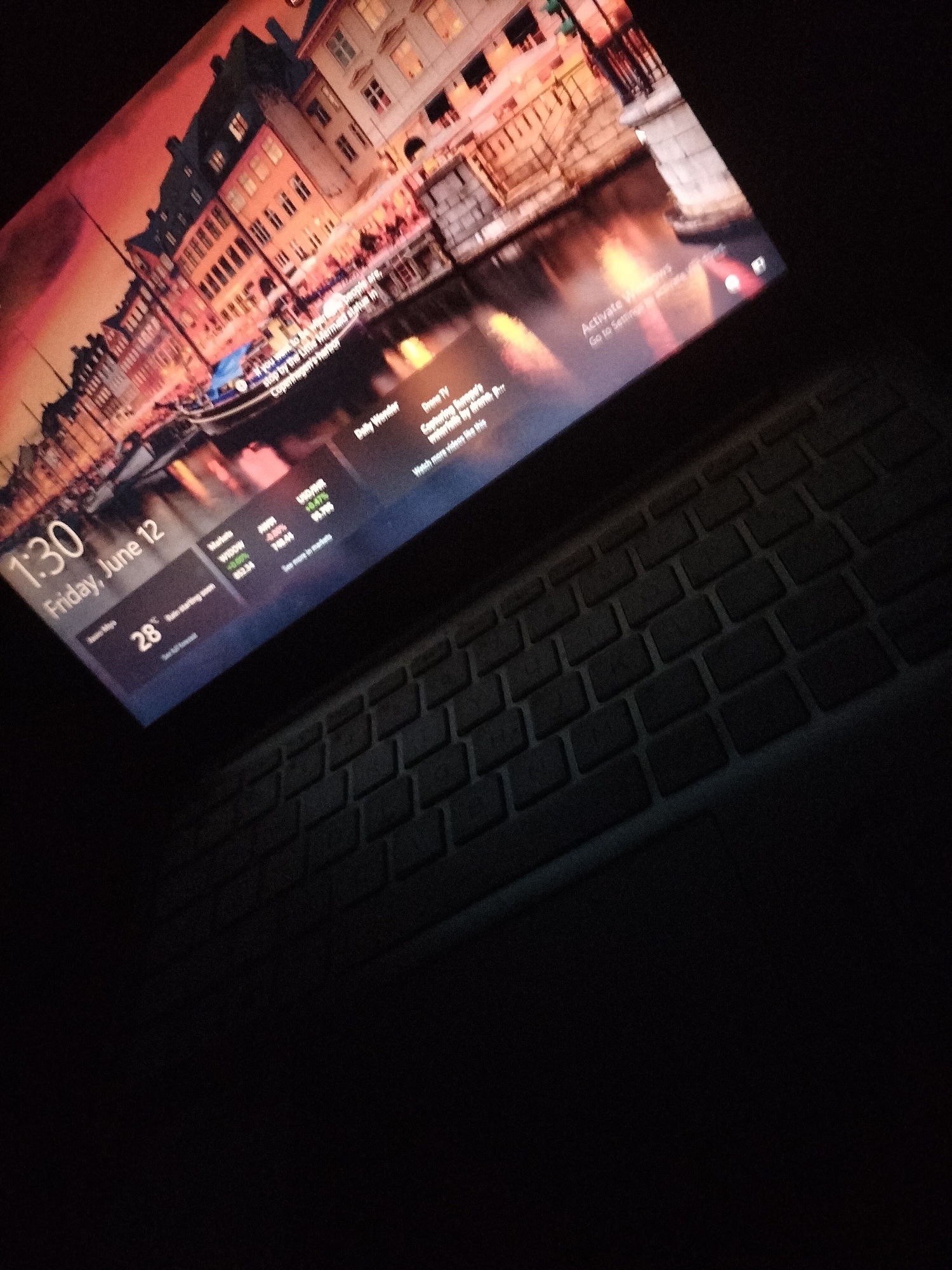 The image shows a laptop with a lock screen that displays a city scene with buildings and a canal. The time is 1:30 on Friday, June 12. There is also a temperature reading of 28 degrees Celsius, indicating a warm day. The laptop is placed on a keyboard, suggesting it is in use. The "Activate Windows" prompt is visible on the screen, indicating the operating system is not activated. The background image appears to be a stock photo of a European city, not Jalingo, Nigeria.