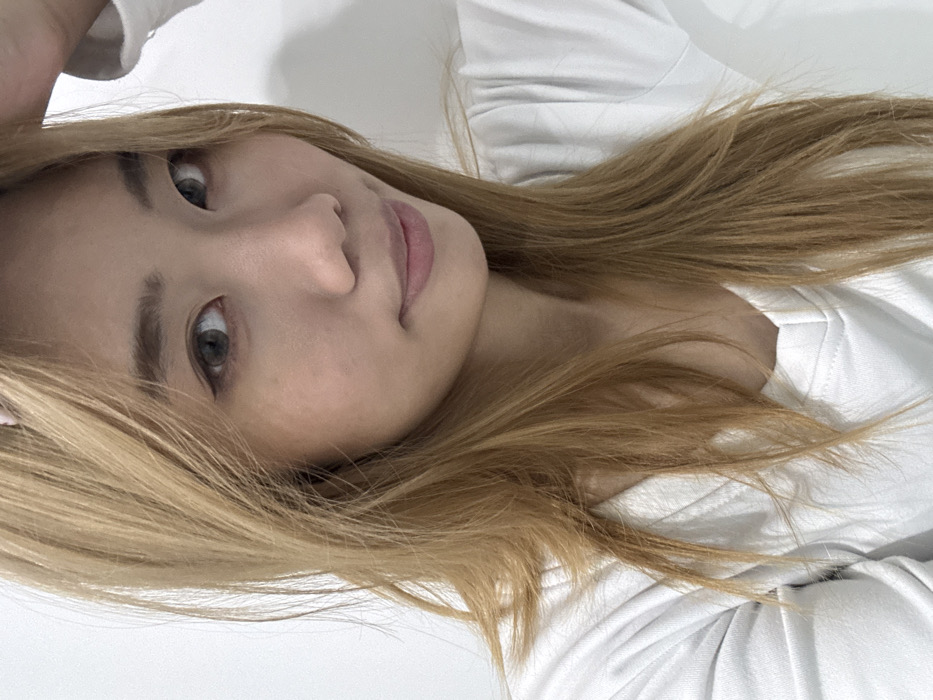 The scene depicts a close-up indoor portrait of an individual, likely female, located in Yuzhong District, China. The subject has fair skin and long, straight to slightly wavy, light brown or dark blonde hair, parted to the viewer's left, extending over her shoulders and partially covering the chest area. A faint pink accessory is partially visible at the top of her head, obscured by hair. Her eyes are open, appearing gray or light brown, with visible dark eyeliner. Eyebrows are dark and well-defined. Her nose is narrow, and her lips are closed, displaying a subtle upward curve at the right corner, colored in a light pink or nude tone. She is wearing a white long-sleeved top with a V-neckline. Her right hand, with fingers partially visible, is raised to the upper right side of her head, touching or positioned near her hair. The subject is gazing directly towards the viewer with a neutral to subtly smiling expression. The background is a plain, undifferentiated white surface, indicating an indoor setting with even lighting.