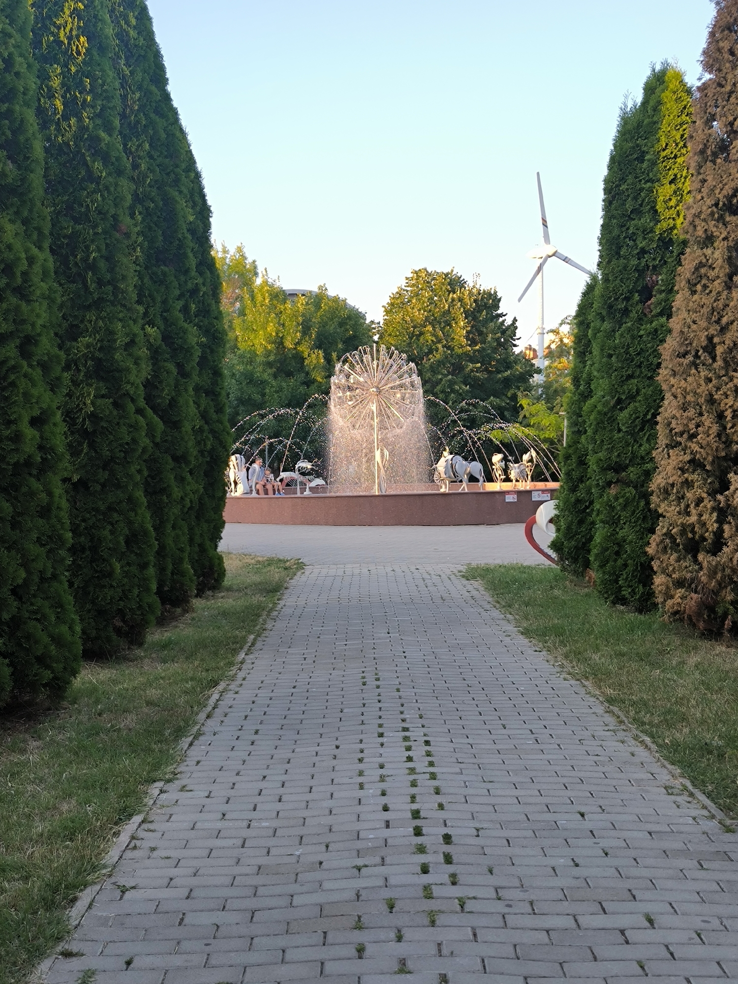 The image depicts a paved pathway leading towards a fountain in a park-like setting. Two rows of tall, green coniferous trees flank the pathway, creating a sense of enclosure. The fountain in the background is active, with water spraying upwards in a spherical pattern. Several people, appearing to be adults and children, are seated around the fountain on a raised platform. In the distance, beyond the fountain and trees, a white wind turbine with three blades is visible against a clear, pale blue sky. The ground surrounding the pathway and fountain is covered in green grass. The scene suggests a public recreational area, likely a park or a public square, in the city of Chișinău, Moldova, given the presence of the wind turbine, which is a known landmark in the city.