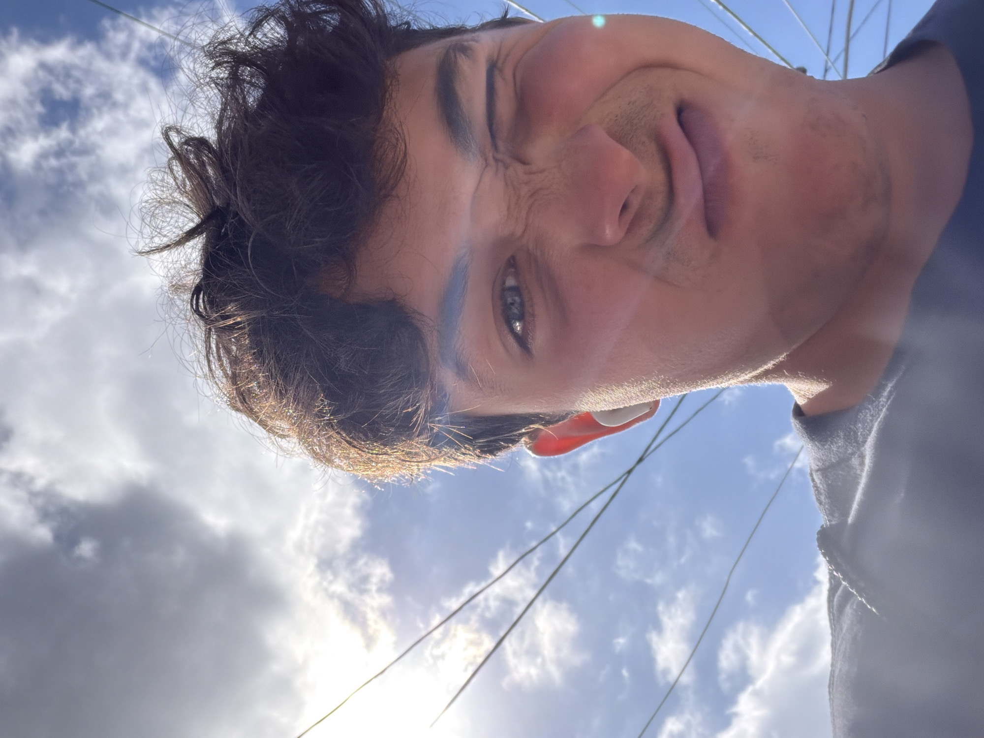 The original image is a low-angle, close-up selfie of a young male with dark, curly hair, taken outdoors against a bright, cloudy sky with visible power lines. He is winking, making a scrunched-face expression, and wearing a dark t-shirt and a white earbud. The sun is shining brightly, creating lens flare and highlighting his hair. The setting appears to be urban, possibly with a slightly coastal feel due to the open sky and power lines, but specific details to confirm Clovelly, Australia are not present in the image itself.