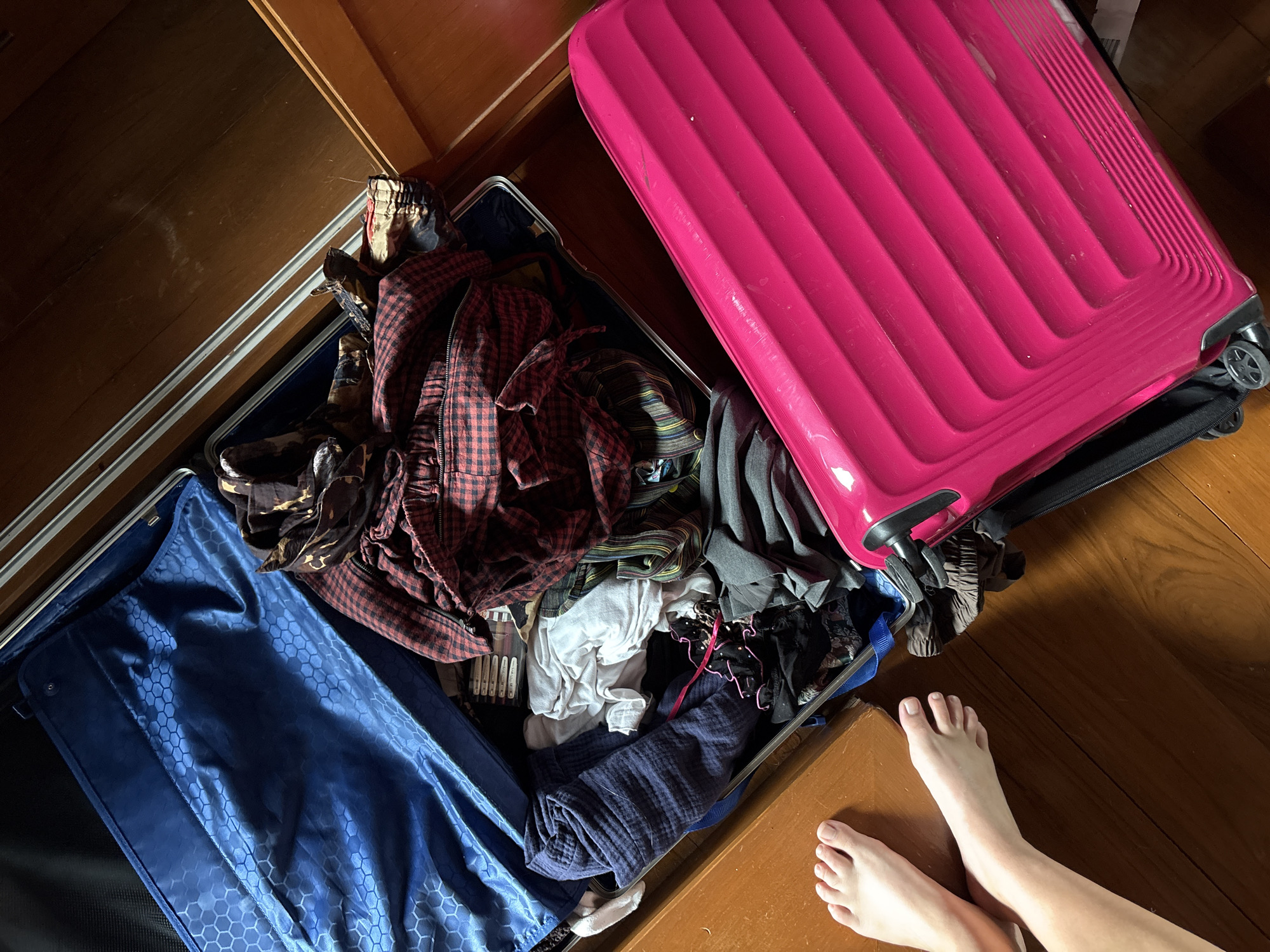 This is a high-angle, top-down shot of two open suitcases on a wooden floor, likely in a bedroom. One suitcase is a vibrant pink, and the other is dark blue. The suitcases are filled with a jumble of clothes, including a red and black plaid shirt, dark pants, and white t-shirts. The scene is lit by natural light, suggesting it's daytime. Two bare feet are visible in the lower right corner of the frame, hinting at a person who is either packing or unpacking. The overall impression is one of disarray, perhaps indicating the chaotic process of preparing for or returning from a trip. The setting appears to be a home, as suggested by the wooden flooring and the presence of personal belongings. There is no visible text in the image.