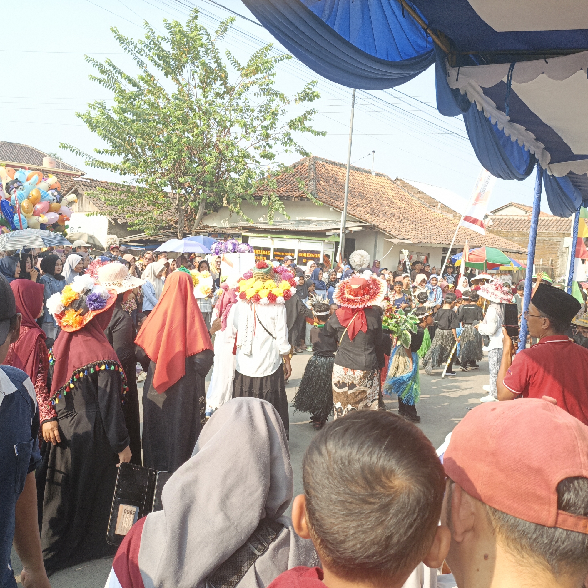 In the city of Tanahbaya, Indonesia, a lively outdoor procession is underway. A group of people, including adults and children, are gathered along a street lined with houses. Many participants are adorned with colorful, oversized hats and traditional attire, suggesting a festive cultural event. In the foreground, several individuals are visible from behind, including a child with dark hair and a person wearing a red cap. A black open wallet, displaying a "5000" denomination card, rests on the ground near a woman in a black outfit with colorful tassels. To the left, a bunch of helium balloons in various colors adds to the celebratory atmosphere. Above, blue and white tents provide shade, and a lush green tree with dense foliage is visible against a clear sky. The overall scene depicts a community engaged in a vibrant celebration.