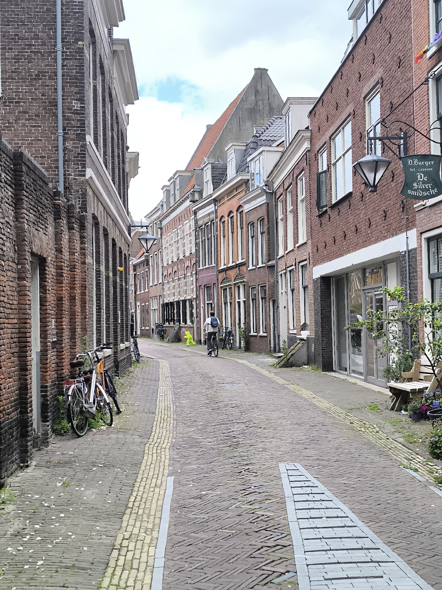 This is an eye-level, medium shot of a narrow, cobblestone street lined with brick buildings in the city of Haarlem, Netherlands. The street slopes slightly upward into the distance. On the left, a tall brick building with arched windows dominates the foreground, with two bicycles parked along its base. Further down the street, more brick buildings with various window styles and rooflines are visible.
A person is riding a bicycle in the middle of the street, heading away from the camera. To the right, another brick building features a hanging sign that reads "D. Burger De Silver smidsche." Next to this building, a wooden bench and potted plants are situated. The sky is overcast with scattered clouds. The overall atmosphere is quiet and residential.