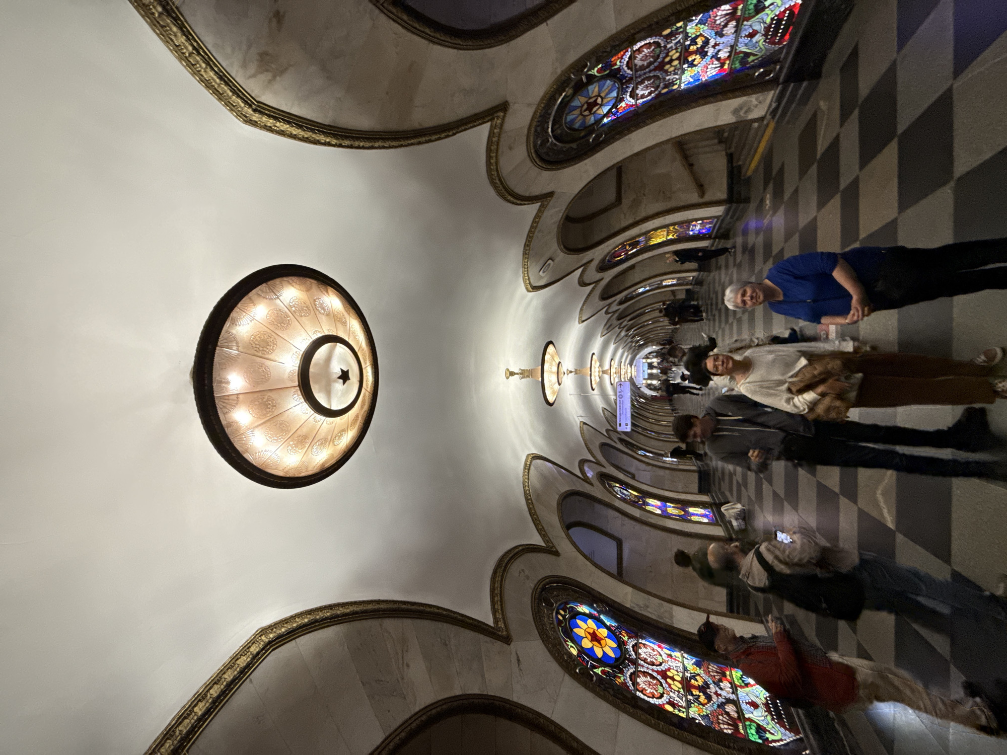 The image captures an ornate subway station with a curved, white vaulted ceiling adorned with gold trim. Large, circular chandeliers with intricate patterns hang from the ceiling. The walls feature arched alcoves with decorative stained-glass windows in vibrant colors. The floor is a checkered pattern of black and white tiles.
In the foreground, four individuals are visible. On the left, a man in a red jacket and a baseball cap faces the camera. Next to him, a person with a backpack looks at their phone. In the center, a woman in a white sweater and brown pants stands looking forward. To her right, a woman in a blue shirt and dark pants also looks forward. Further down the platform, other people are seen walking and standing. The overall scene depicts a well-lit, grand public space.