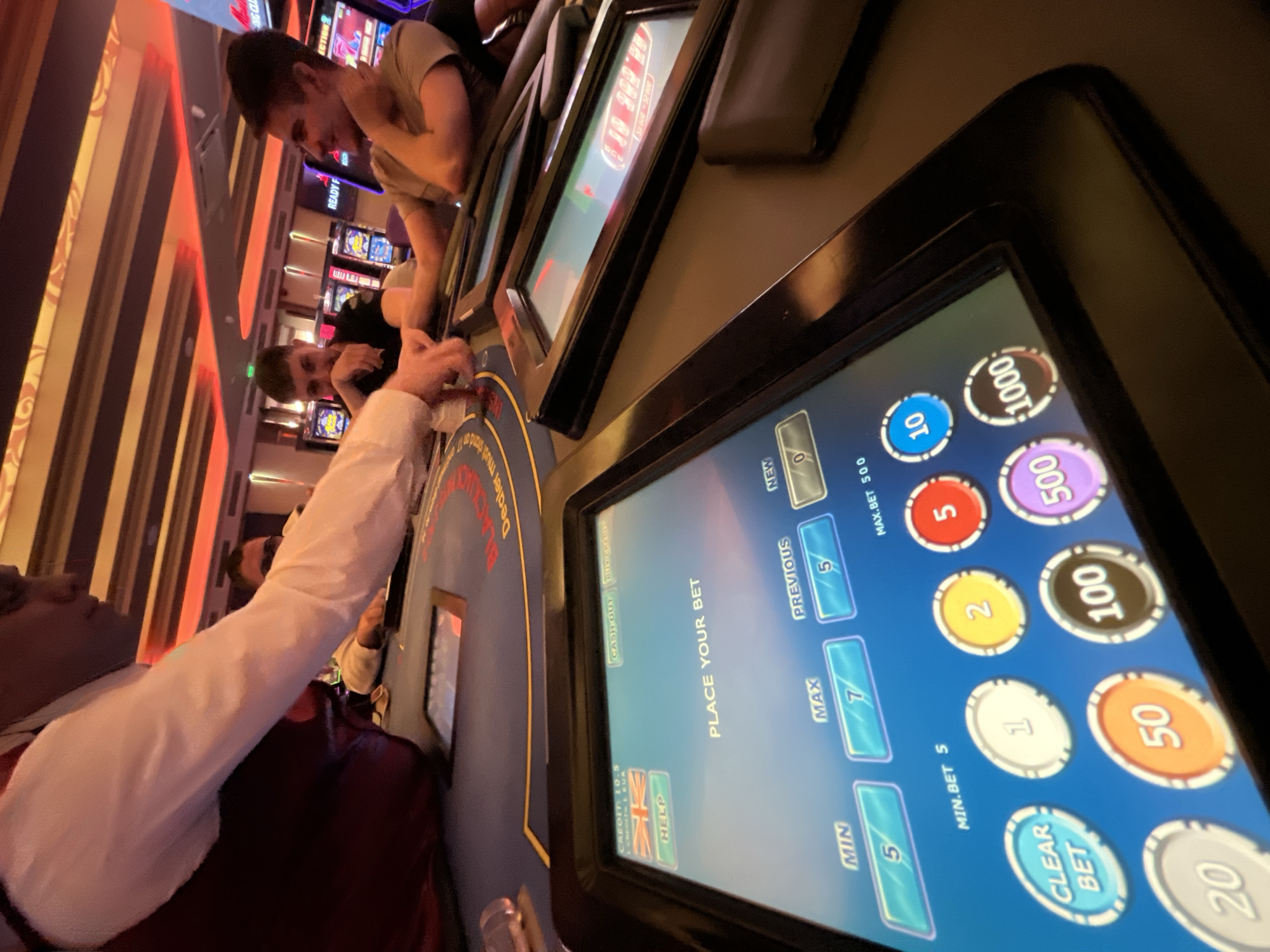 The image depicts a scene inside a casino in Burgas, Bulgaria. Two people are visible at an electronic blackjack table. A man in a white long-sleeved shirt is interacting with the touchscreen interface of the table, which displays options for placing bets, setting minimum and maximum bet amounts, and chip values. To the right, the screen shows values like 5, 10, 20, 50, 100, 500, and 1000. To the left is a screen showing READY F and other slot-machine-related information. The words "DEALER must stand on 17" are also visible on the table. A man in a t-shirt is leaning on the blackjack table, observing. The casino environment is illuminated with artificial lighting and the interior details include striped and decorated walls.