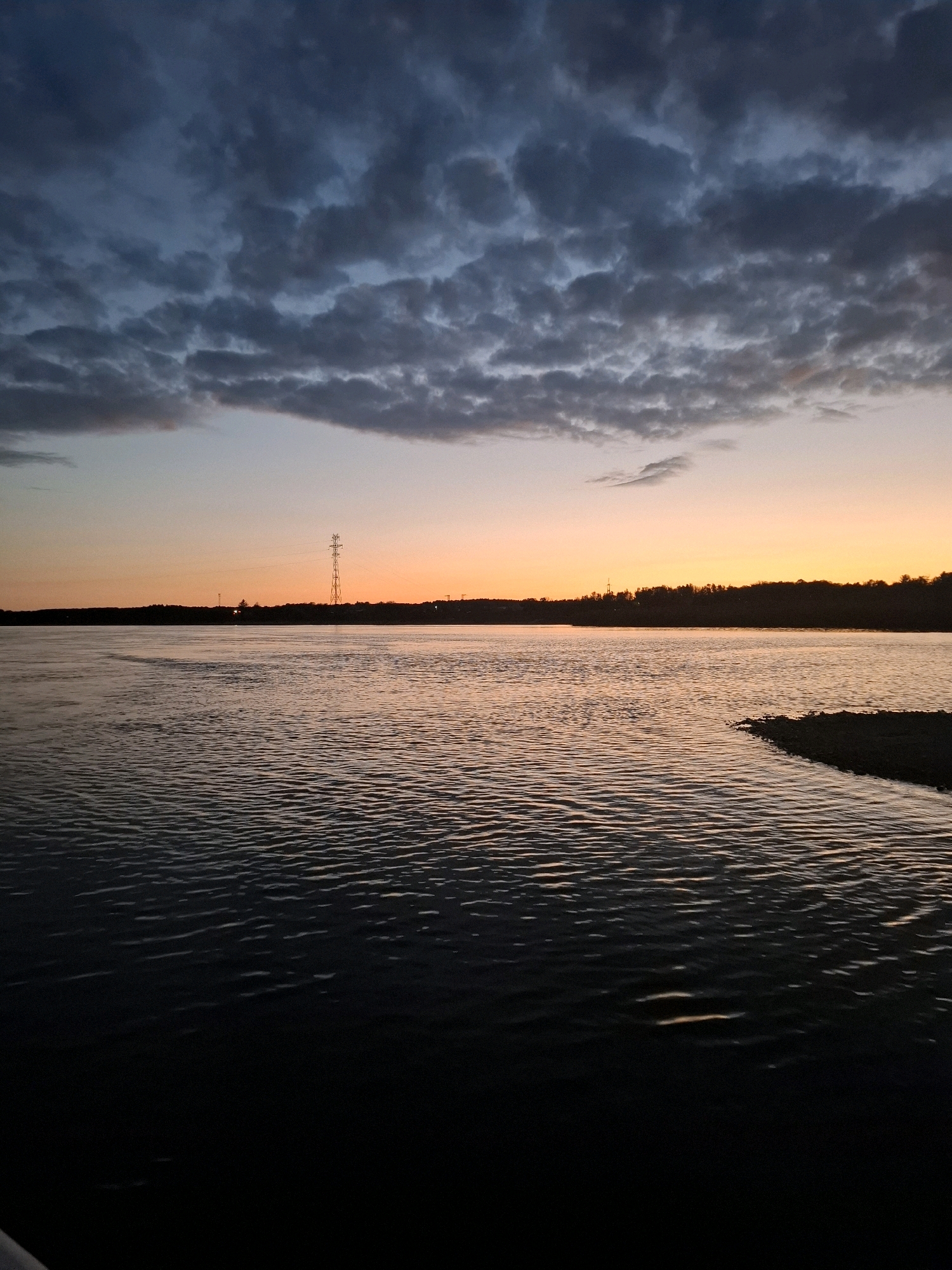 The image depicts a dusky sky over a body of water. Dark, brooding clouds fill the upper portion of the frame, transitioning to a soft gradient of orange and yellow near the horizon, indicating either sunrise or sunset. A silhouetted tree line runs along the far bank of the water, with a prominent electrical tower visible against the lighter sky. The water in the foreground is dark and rippled, reflecting the muted colors of the sky. The overall scene is serene and slightly melancholic, with no people or visible activity. The location is identified as being in the city of Bardagon, Russia, though no specific landmarks or features within the city itself are identifiable in the image.