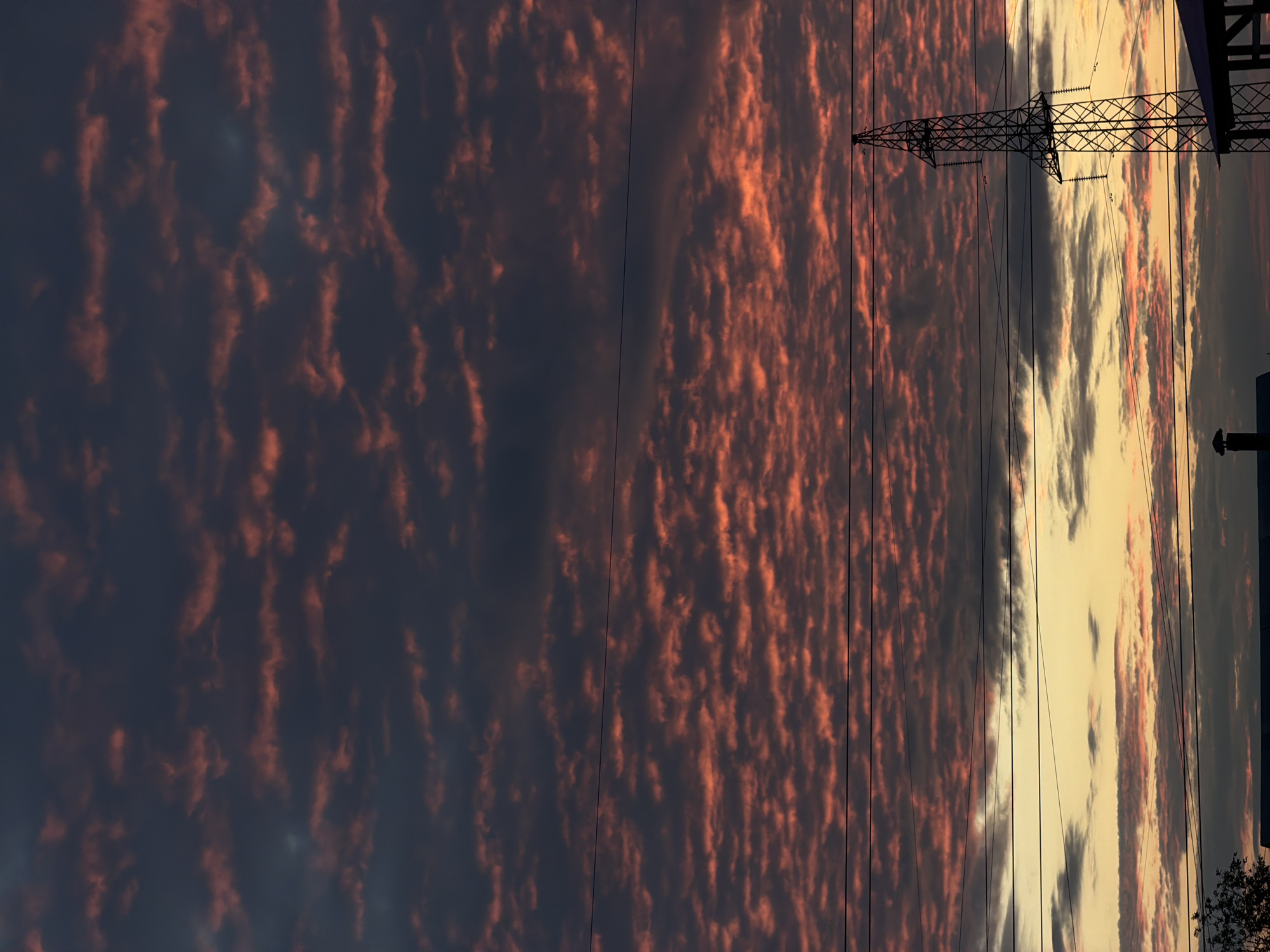 The image depicts a dramatic sunset in Chaikovsky, Russia, with a sky filled with dark, textured clouds tinged with vibrant coral and orange hues. These clouds are arranged in a rippled pattern, creating a visually striking spectacle. Several power lines stretch horizontally across the frame, dividing the sky and adding a man-made element to the natural beauty. In the lower right quadrant, a tall, intricate electricity pylon stands in silhouette against the colorful sky, its lattice structure clearly defined. Below the pylon, the dark silhouettes of rooftops and a chimney are visible, suggesting a residential or industrial area. The overall scene captures a moment of natural grandeur juxtaposed with the utilitarian presence of human infrastructure at dusk.
