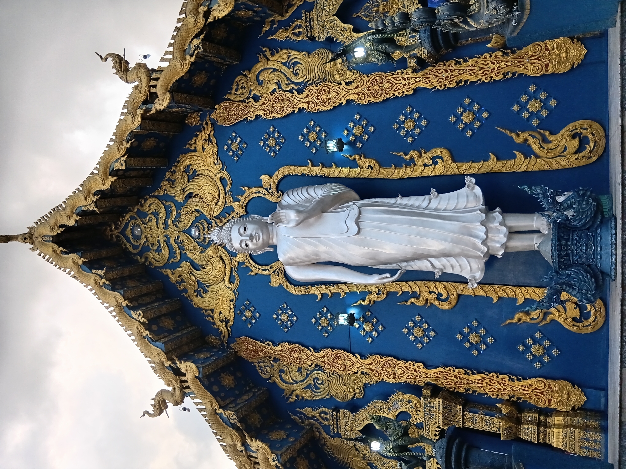 The image shows the Wat Rong Suea Ten, also known as the "Blue Temple," located in Chiang Rai City Municipality, Thailand. The temple is characterized by its vibrant blue facade adorned with intricate gold carvings and decorative elements. Prominently featured in the foreground is a tall, white statue of Buddha standing with his right hand raised in a gesture of blessing. The Buddha statue is positioned in front of the main temple building, which has a steeply pitched, multi-tiered roof decorated with golden mythical creatures, possibly Naga serpents, and a golden finial. The blue walls of the temple are decorated with golden floral motifs and diamond-shaped patterns. Two lanterns are visible on the walls, one to the left and one to the right of the Buddha statue. Dark, ornate statues of mythical creatures flank the entrance, adding to the grandeur of the temple's exterior. The sky above is overcast, suggesting a cloudy day.