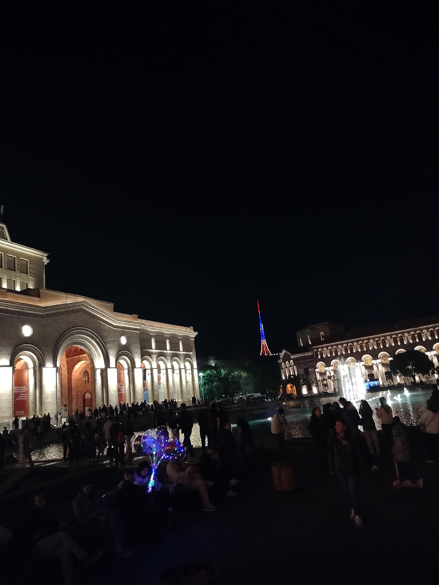 The image shows a night scene at Republic Square in Yerevan, Armenia. On the left and right sides are large buildings with arched facades illuminated with warm light. The building on the left appears to be the National Gallery of Armenia. A fountain is in the center-right of the image with water jets lit up. Many people are gathered in the square, some sitting and some walking around. There are also some glowing balloons being held in the crowd. In the background, between the two buildings, a tall tower is lit up with the colors of the Armenian flag: red, blue, and orange. The sky is dark and clear above.
