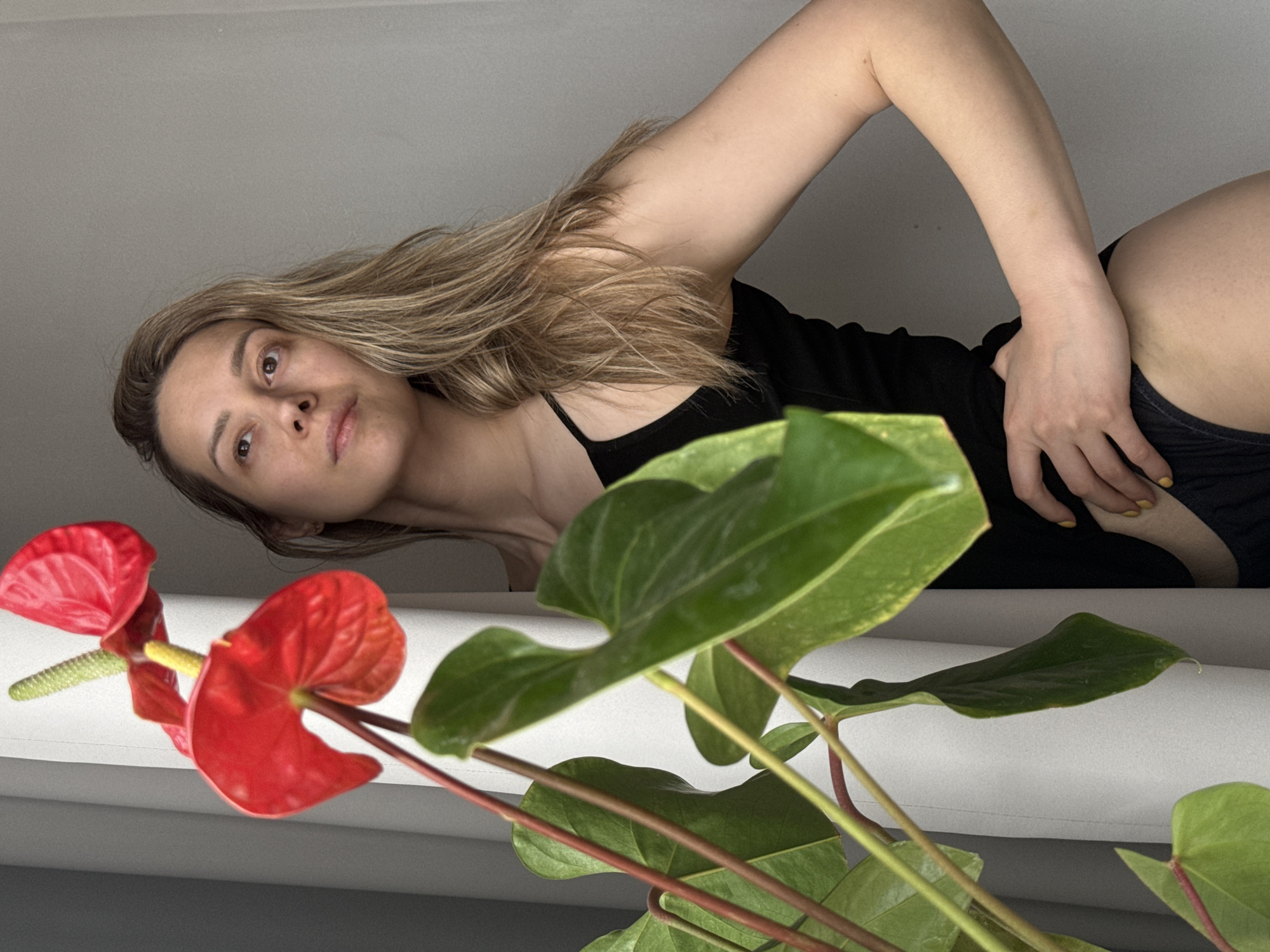 A medium shot, taken indoors, shows a fair-skinned woman with light brown hair. She is wearing a black tank top and black shorts, and is positioned behind a plant with red flowers and large green leaves. The background is a plain wall. Her left hand is near her hip, with her fingers gently touching her shorts. The photo is taken from an unusual angle. The image does not contain enough information to ascertain a location in Chaikovsky, Russia.