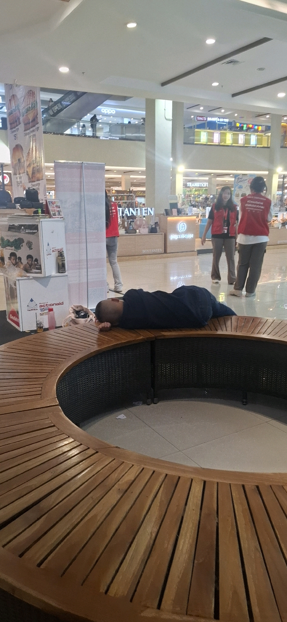 A man is sleeping on a wooden bench in a mall. He is wearing a dark blue shirt and dark pants. The bench is curved and made of wood slats. Behind him, there are two women wearing red vests and grey pants, suggesting they might be employees. One of the women is walking, and the other is standing. In the background, there are shops and escalators. The mall appears to be well-lit and spacious. The man's head is resting on the edge of the bench, and his body is partially covered by his shirt. The overall scene suggests a public space where people might rest or wait. This scene likely takes place in a mall in Surakarta, Indonesia, given the context.