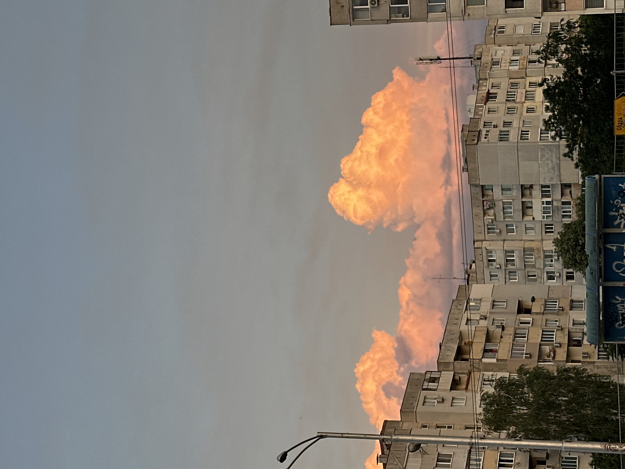 The image depicts a sunset scene over a residential area in Sofia, Bulgaria, characterized by apartment buildings and dramatic, illuminated clouds. The sky is a gradient of soft blues and grays, transitioning to a warm, peachy orange hue where the clouds catch the sunlight. These clouds are large and voluminous, appearing to glow from within.
In the foreground, several multi-story apartment buildings dominate the landscape. They are constructed in a Brutalist or Soviet-era architectural style, with concrete facades and numerous repetitive windows. Many of these windows are visible, some with air conditioning units protruding from them. The buildings are densely packed, suggesting an urban environment. Trees, some appearing quite leafy and green, are interspersed between the buildings and in front of some of them.
A street light with two lamps stands prominently on the left side of the image, its metallic structure stark against the sky and buildings. Power lines are also visible, crisscrossing the frame above the apartment blocks. A blue structure with graffiti on it is partially visible at the bottom of the image, possibly a bus stop or kiosk. A yellow sign with text in Bulgarian is also visible in the lower right corner, indicating signage for directions. The overall impression is of an ordinary urban evening, with the natural beauty of the sunset contrasting with the utilitarian architecture of the city.