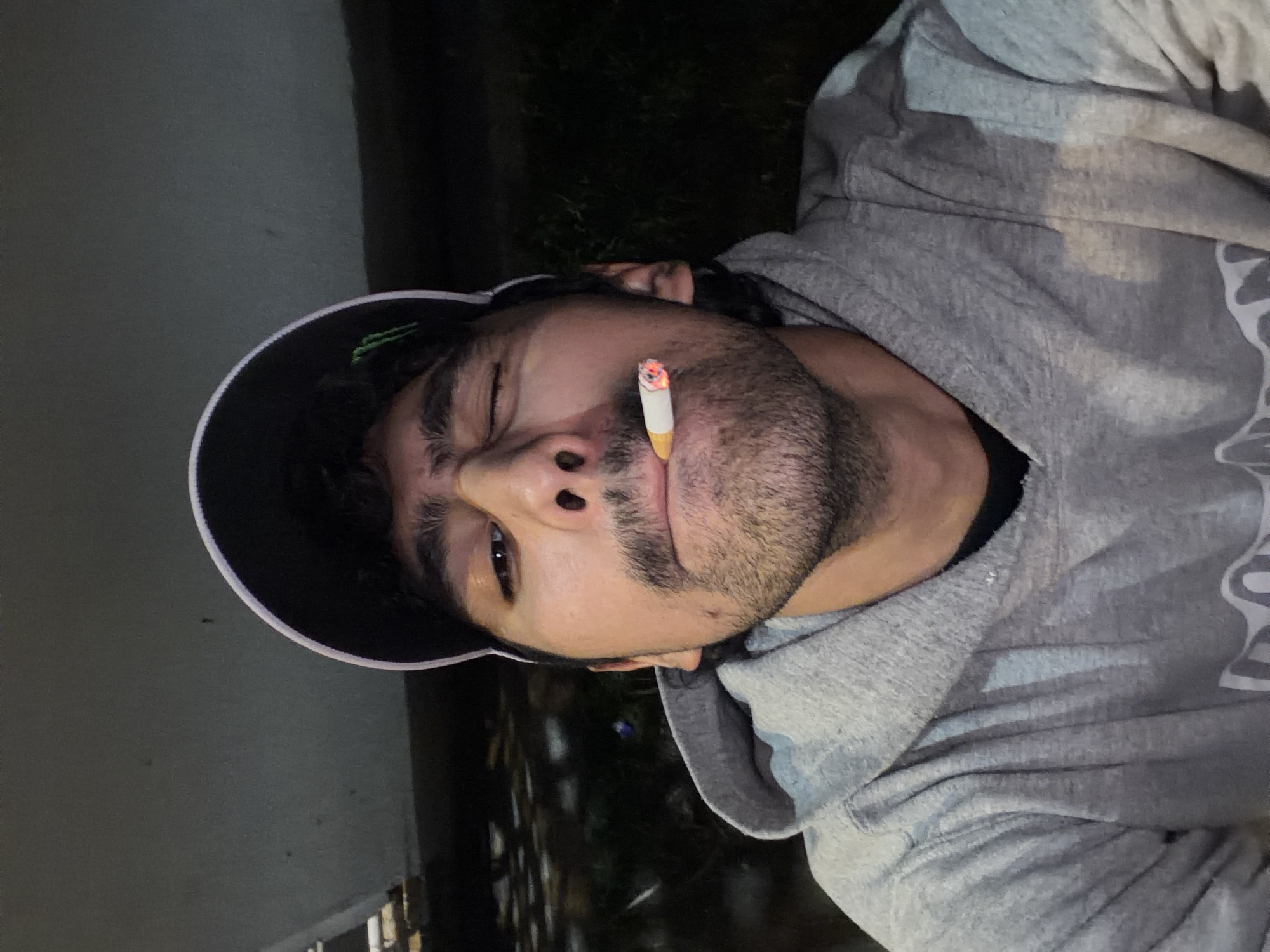 A man with dark hair and a beard lies on his side outdoors. He is wearing a gray hoodie and a black baseball cap with a white trim, and he is smoking a lit cigarette held in his mouth. The background includes a dark area of vegetation and what appears to be a building wall. The location may be the city of San Andrés Cholula, Mexico.