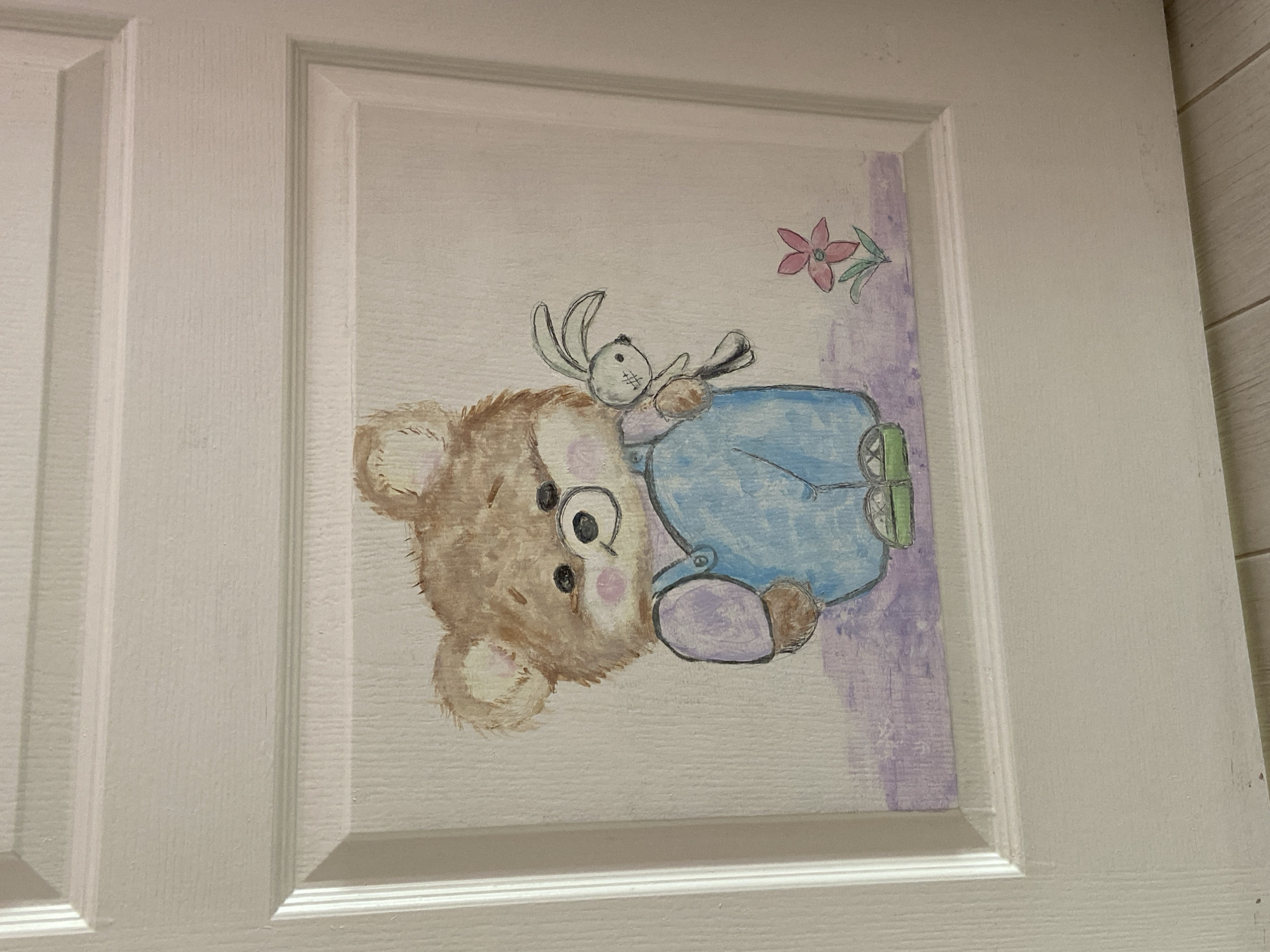 The image shows a painted panel on a white door. The artwork depicts a brown teddy bear wearing blue overalls, lying on its side. A small white rabbit is perched on the bear's chest. A pink flower is located above the bear. The door appears to be indoors. The exact location in the city of Unknown, Unknown is not determined from the image.