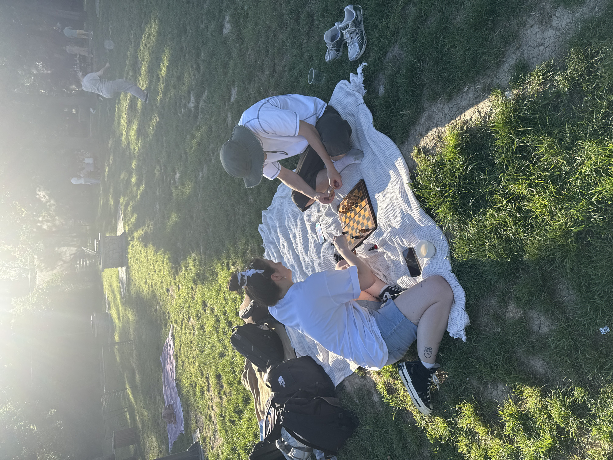 Two people are playing chess on a blanket in a grassy area. One person is kneeling and wearing a hat, shorts, and a white t-shirt. The other person is sitting and wearing denim shorts, a white t-shirt, and black sneakers. There are shoes, backpacks, and a cell phone on the blanket. The location appears to be a park or grassy area within the city of Cukarica Urban Municipality, Serbia. There are trees and other people in the background. The sun is shining.