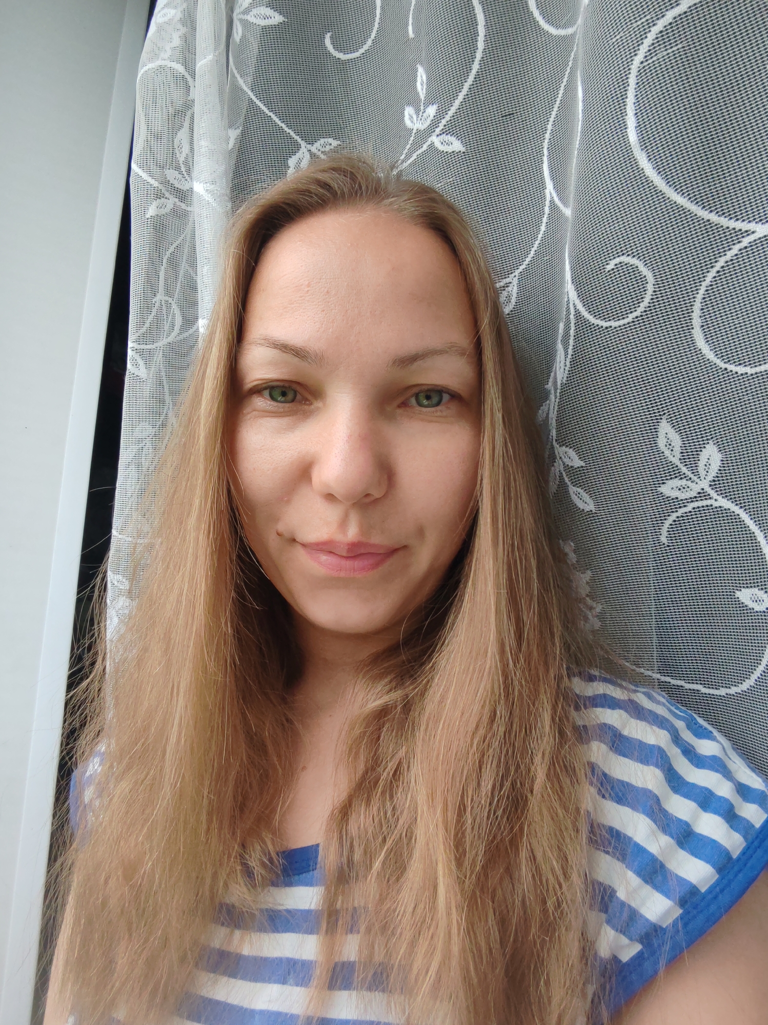 A blonde woman with green eyes and fair skin is pictured in a close-up, head-and-shoulders portrait. She has long, straight, light brown hair that falls around her shoulders. She is wearing a blue and white striped t-shirt. Behind her, a white lace curtain with a floral pattern is visible, obscuring a window or wall. The woman is looking directly at the camera with a slight smile. The setting appears to be indoors, near a window. The image does not provide any specific indicators of a location in the city of Чухлэм, Russia.