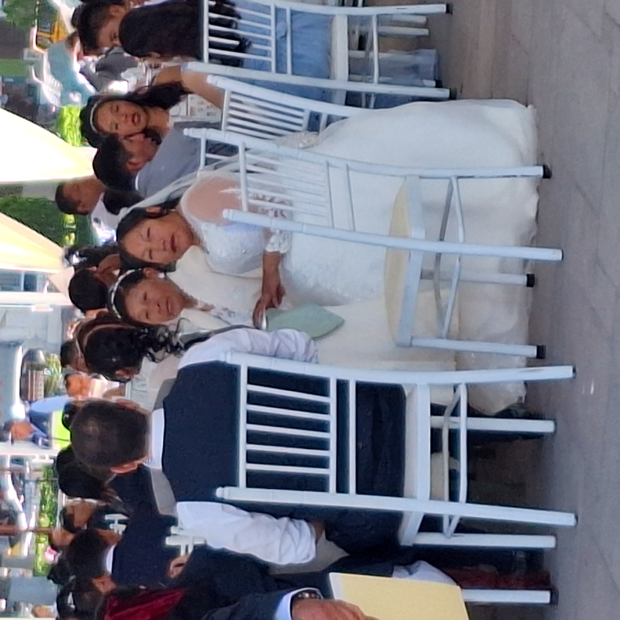 The image shows a group of people seated at an outdoor event, possibly a wedding, in Cerro Colorado, Peru. Several individuals are wearing formal attire. Two women are in white gowns, suggesting they are brides. A man in the foreground wears a suit and holds a yellow folder. White chairs are prominently placed between the camera and the subjects. The environment appears to be a paved area.