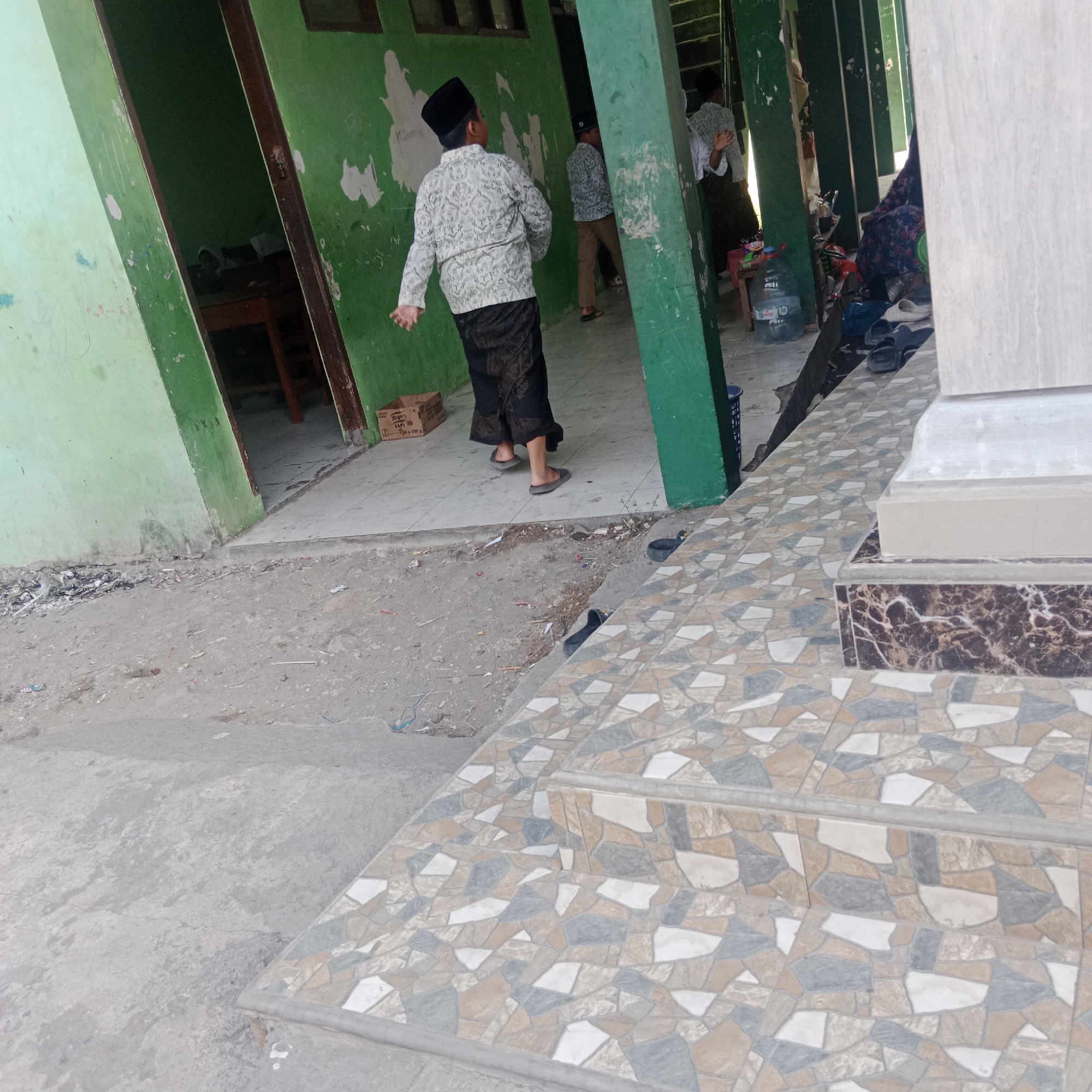 In a street scene in Tegowanu Kulon, Indonesia, two boys in traditional Islamic attire stand near a green building. The boy in the foreground wears a white patterned shirt and a black peci hat, with a patterned sarong. He is captured mid-motion, with his left arm extended. Another boy, also in similar attire, is visible further down the walkway. The building has peeling green paint and an open doorway leading to a dark interior with a wooden table. The ground is a mix of concrete and dirt, with scattered debris. In the foreground, a tiled pathway with a mosaic of geometric shapes ascends, with a marble-patterned step. Several pairs of sandals are visible on the ground near the building's entrance. A large bottle of water and a blue basket are also present.