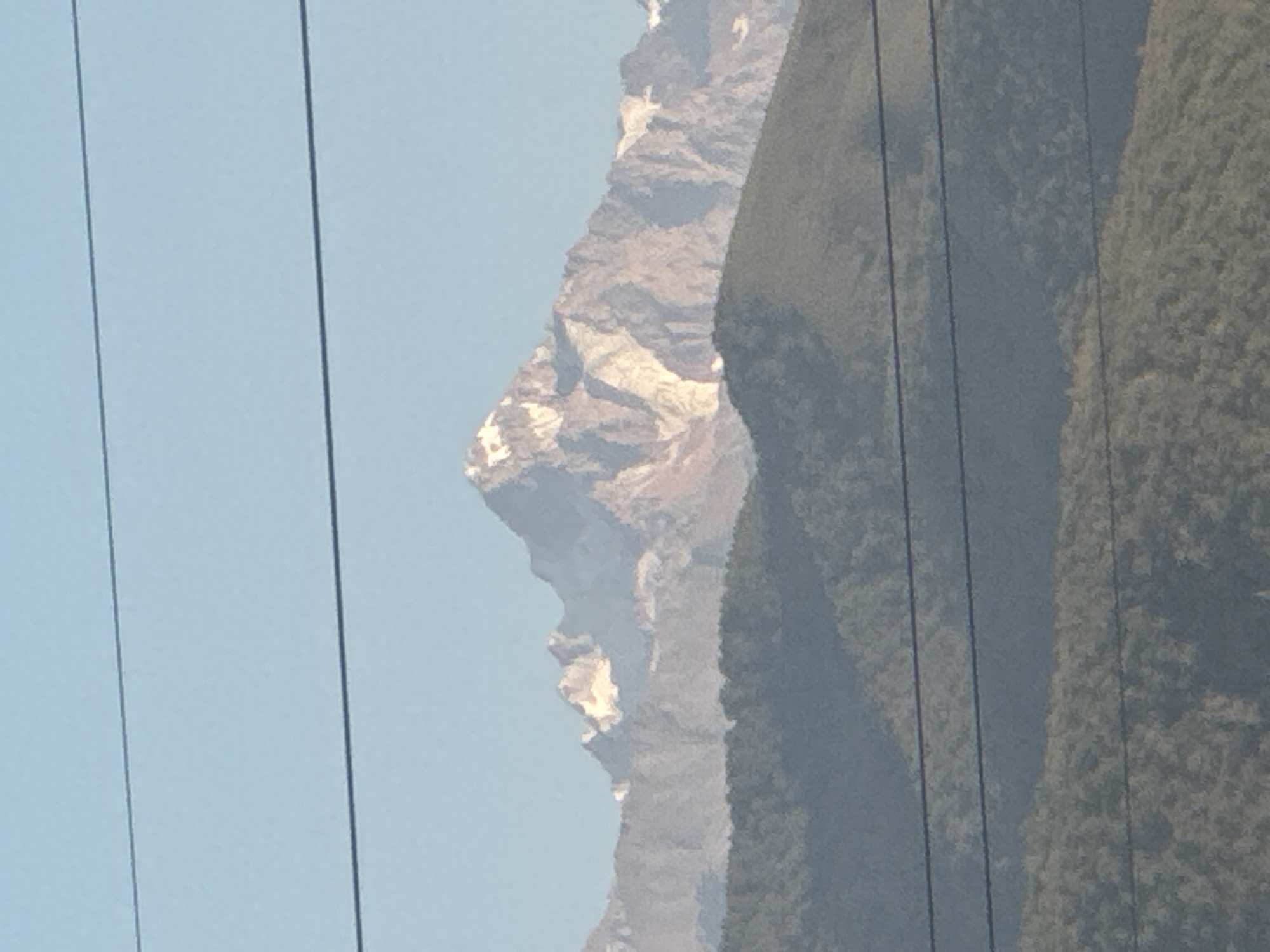 The image shows a distant view of snow-capped mountains under a clear blue sky. In the foreground, there are blurred, dark horizontal lines that appear to be power lines. The mountains are rugged and show significant snow cover, particularly on the higher peaks. The lower slopes of the mountains are covered in vegetation, appearing as dark green, undulating shapes. The overall scene suggests a natural, mountainous landscape. No people, animals, or specific structures are visible in the image. The quality of the image is somewhat degraded, with a lack of sharp detail, possibly due to distance, atmospheric conditions, or the camera used. The scene does not provide any specific visual cues to confirm the location as Tuzdybastau, Kazakhstan, but it is consistent with a mountainous environment that might be found in such a region.