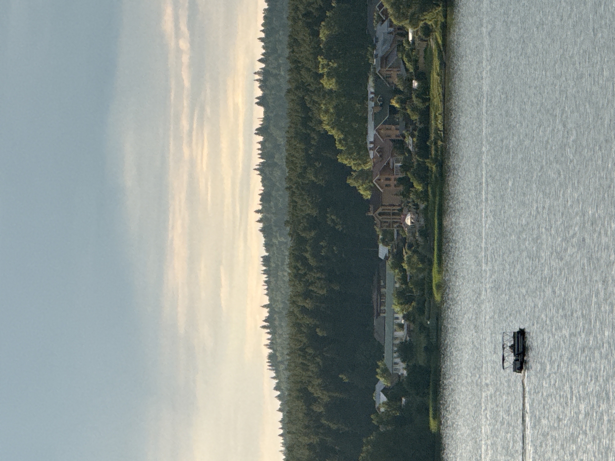 A pontoon boat with a canopy is moving across a body of water. A row of houses and trees line the shore in the background, with a dense forest beyond. The sky above is a pale blue with wispy clouds. The image appears to be taken from a distance, possibly from another boat or a higher vantage point. The location is Votkinsk, Russia.