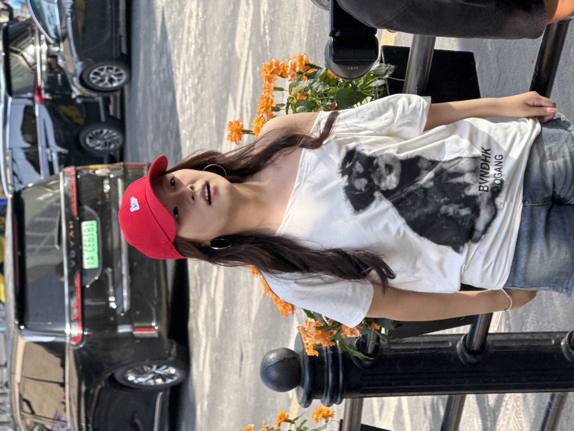 In Yuzhong District, China, a young East Asian female stands outdoors, wearing a red baseball cap and a white off-the-shoulder t-shirt with a black and white graphic of a dog and text at the bottom. She also wears denim shorts and a silver bracelet. She is looking towards the right with her mouth slightly open. Behind her, several dark-colored SUVs are parked. Orange flowers are in planters to her left. A black railing is in the foreground.