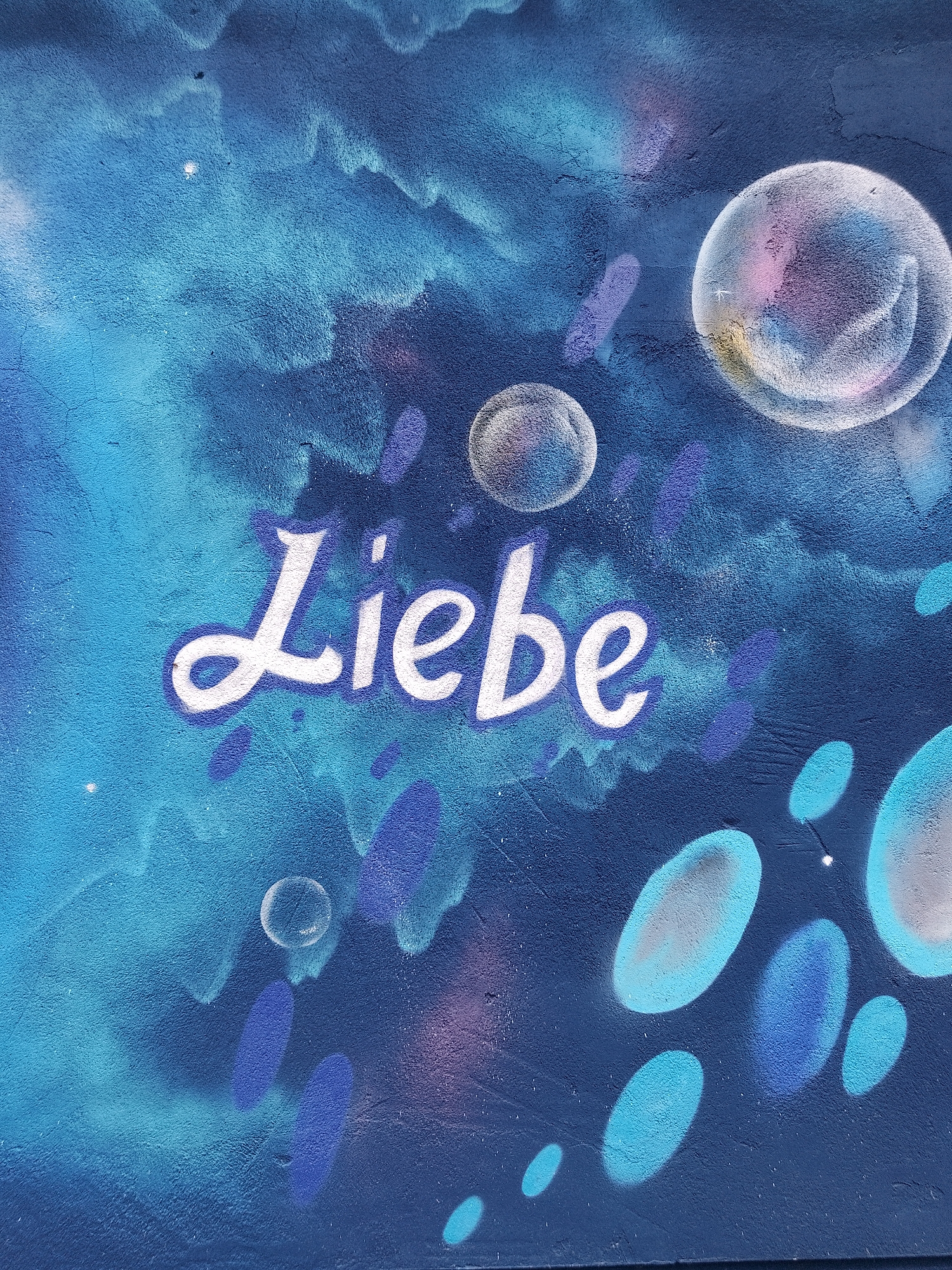 The image shows a blue wall mural in Unknown, Unknown. The mural depicts a cloudy blue background with bubbles of various sizes. The word "Liebe," which means love in German, is painted in white script in the center of the image.