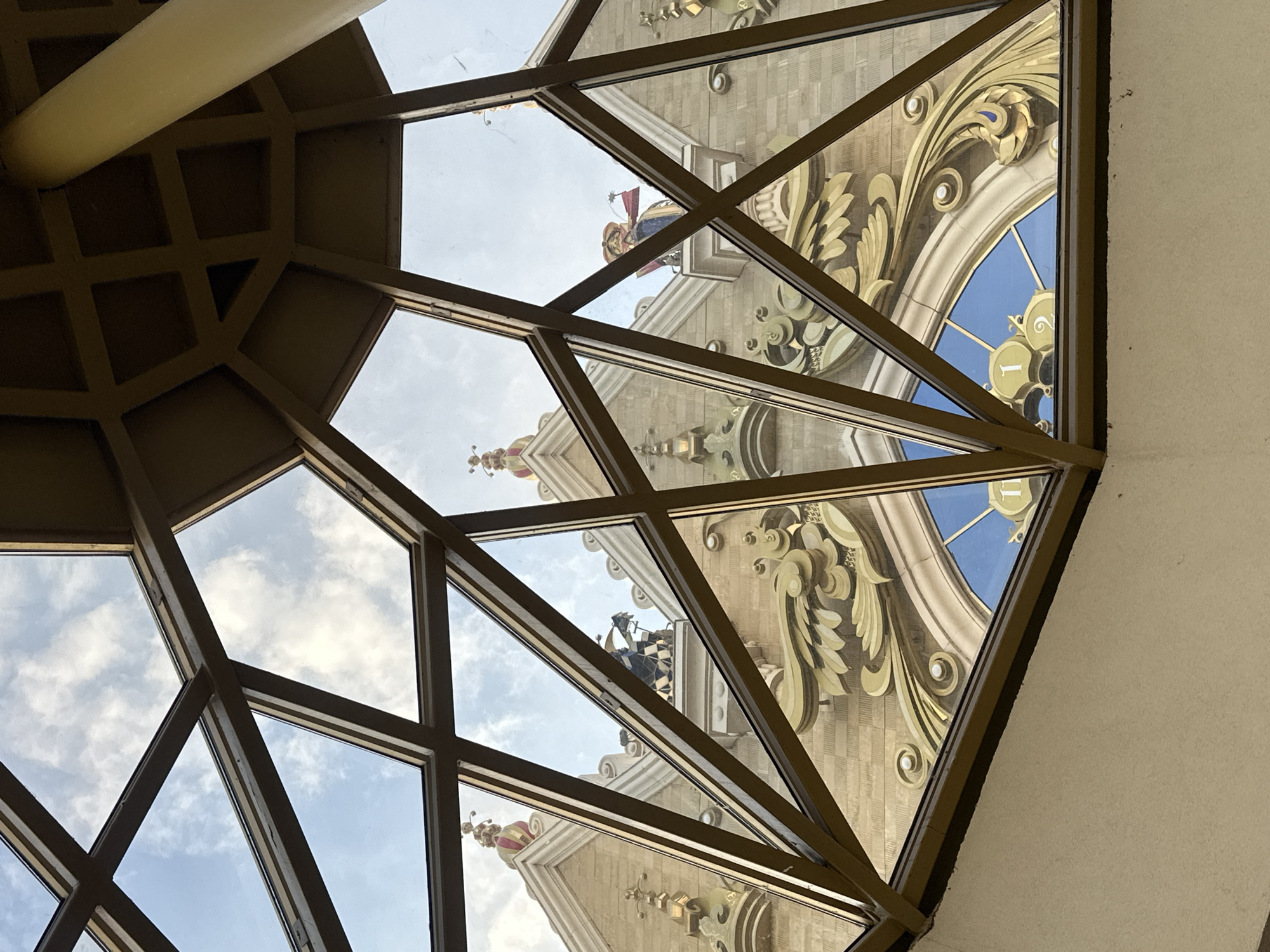 The image shows an upward view through a geometric window in Kazan, Russia. The window's structure is a network of brown metal bars forming triangular panes of glass. Through the glass, a section of an ornate building façade is visible. The building has gold floral decorations, blue accents, and statues. A clock face can be seen on the building. The sky is visible through other panes, with scattered clouds.