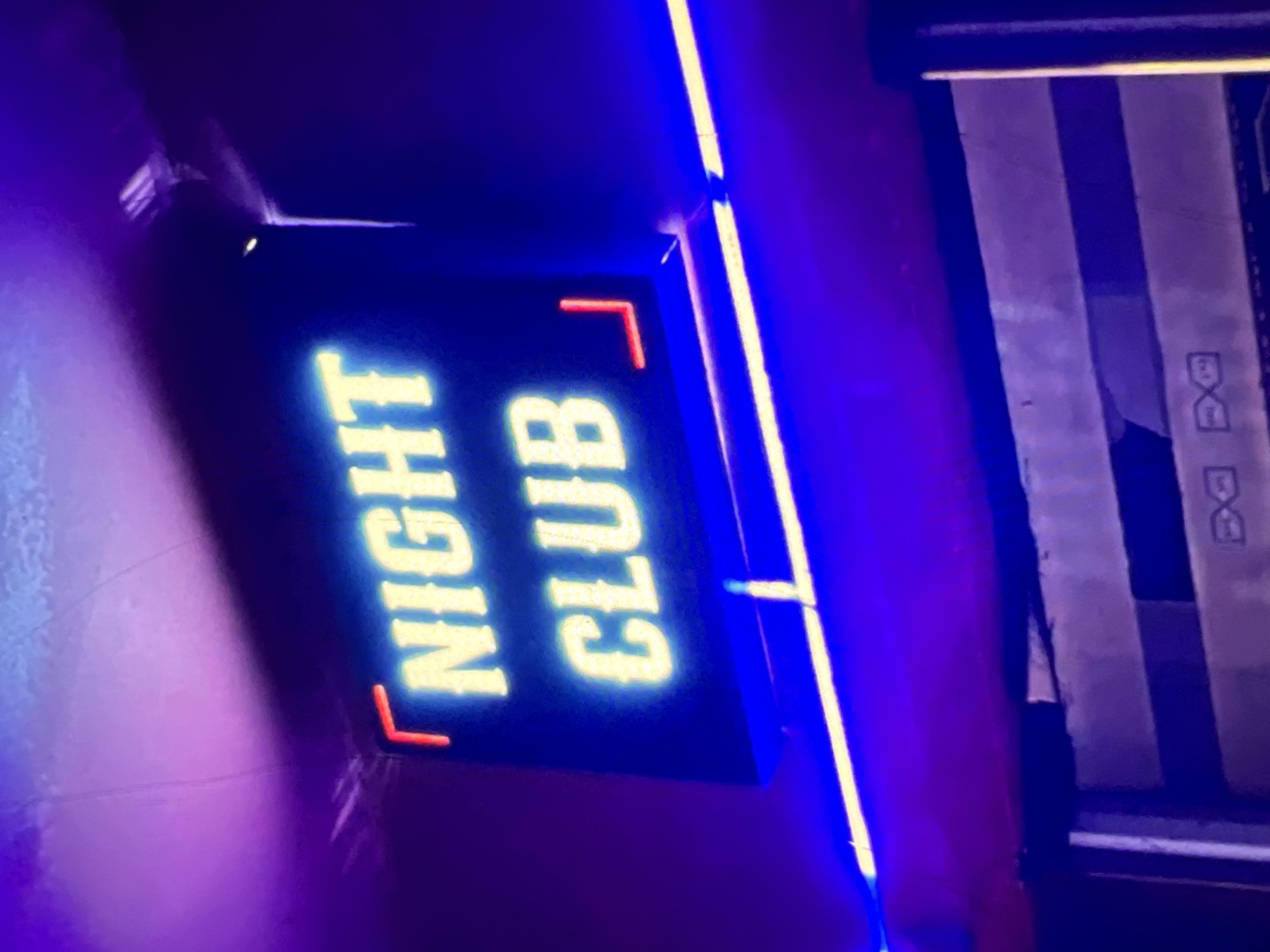 The image shows a neon sign that reads "NIGHT CLUB" in bright yellow lettering against a dark background. The sign is illuminated by purple and blue lighting, creating a vibrant and atmospheric effect. The sign appears to be mounted on a wall or ceiling, and a bright blue neon light strip runs horizontally below it. The lower part of the image shows a glimpse of what looks like a stage or a bar area, with some grey and dark panels and a faint pattern that could be part of a logo or design. There are no people visible in the image. Based on the visual style and the type of establishment, this sign is likely located in a nightlife district within Lagos, Nigeria.