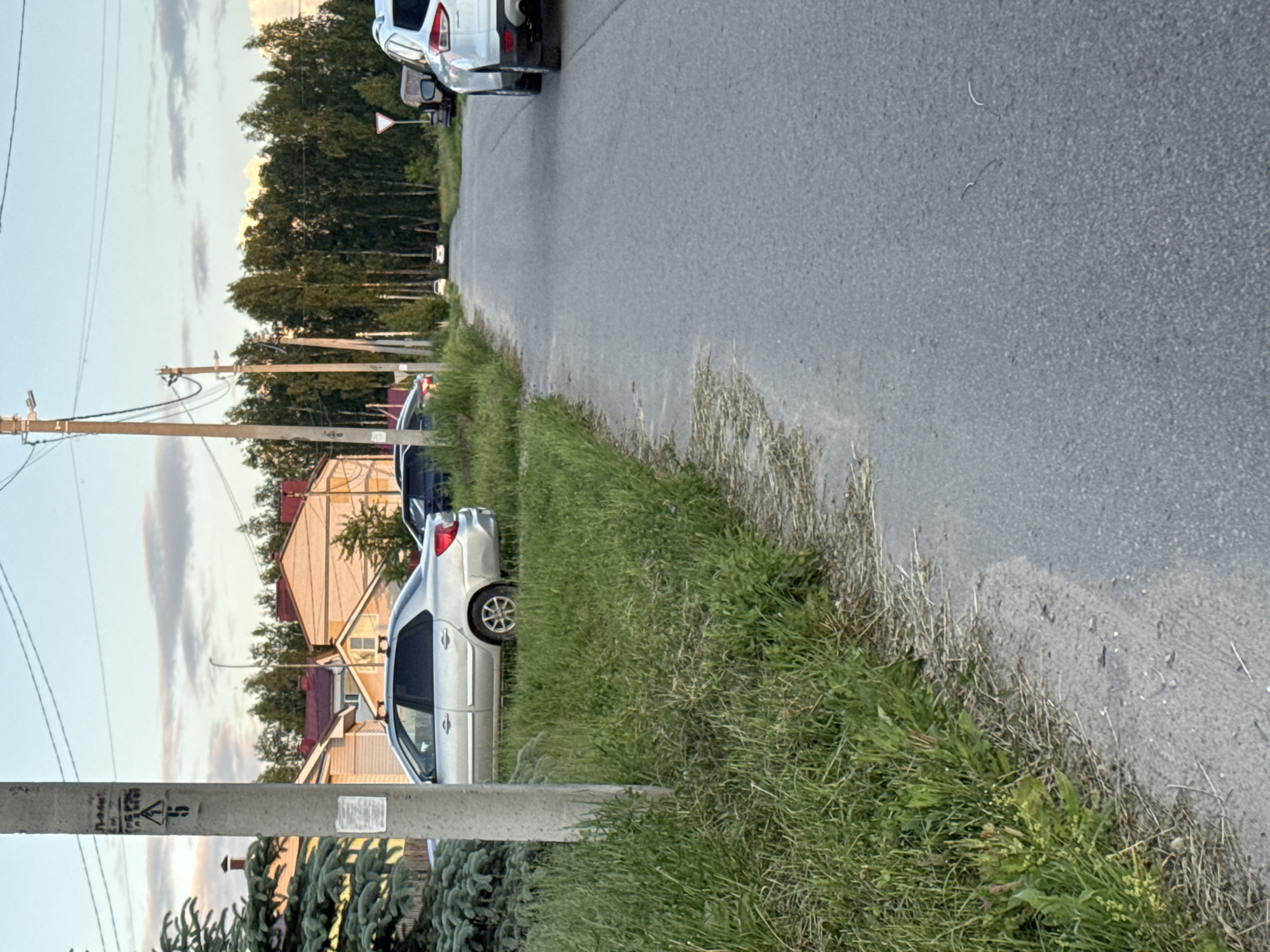The image depicts a street scene in Krasnye Baki, Russia. The viewpoint is from the side of a paved road, with a grassy embankment to the left. Several cars are parked along the side of the road, including a silver sedan, a dark-colored sedan, and a white SUV. Residential houses with red roofs are visible in the background, as well as a line of tall trees. Utility poles with wires are present along the street. The sky is partly cloudy with a soft, diffused light, suggesting either early morning or late afternoon. The overall impression is of a quiet, suburban street.