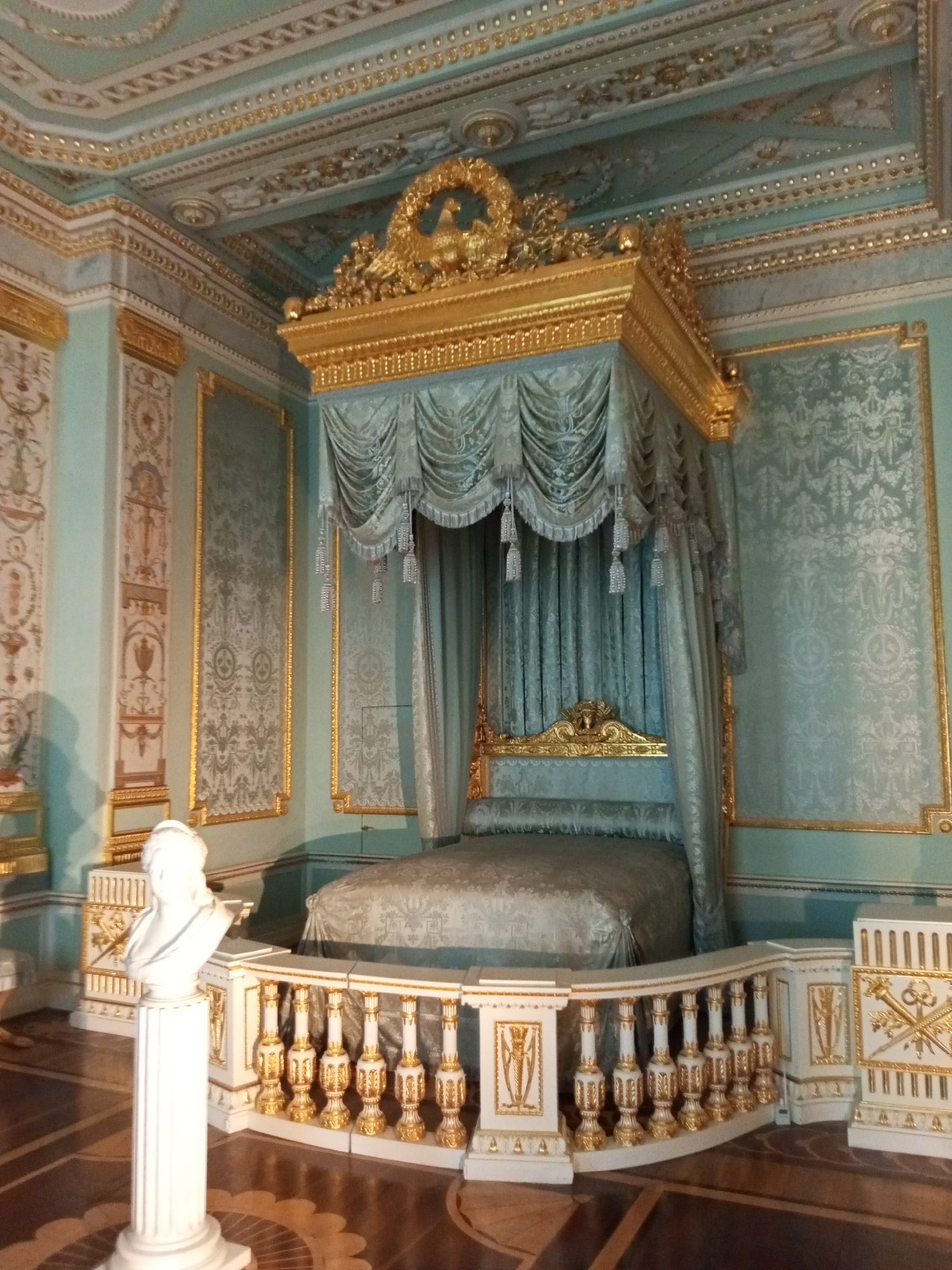 The image depicts a luxurious bedroom in an opulent palace. The focal point is a four-poster bed with a gold frame and light blue drapes. The walls are adorned with patterned light blue wallpaper and framed with gold trim. The ceiling is ornate, featuring intricate gold details. A white bust stands on a pedestal near the bed, which is surrounded by a white and gold railing. The floor is made of dark wood with a decorative pattern. The overall impression is one of wealth and grandeur. The location is Unknown, Unknown.