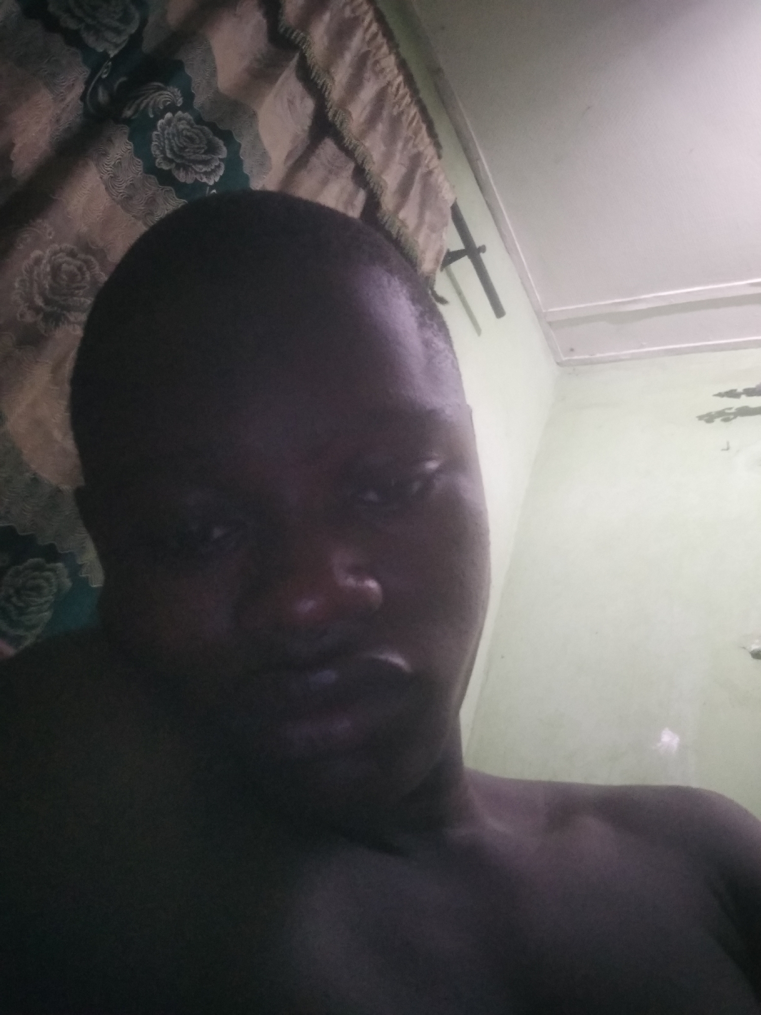 A dim, close-up photograph captures an individual with dark skin tone, likely a selfie. The person's face is partially obscured by shadows, with some illumination on their eyes, nose, and mouth, revealing a neutral or slightly serious expression as they look towards the camera. Their short hair is visible, and the upper part of their body, including shoulders and upper chest, is also in the frame, appearing dark due to the low light. They seem to be shirtless or wearing dark, form-fitting clothing.
The background of the indoor setting consists of a patterned curtain on the left, featuring dark green fabric with light-colored floral (rose) designs and a fringed edge. To the right and above the person, a pale green wall with some visible marks or discoloration meets a white ceiling. A thin, dark, horizontal fixture, possibly a curtain rod or support, is mounted on the wall. The overall lighting is low, creating a moody atmosphere and emphasizing the shadows on the subject. There is no discernible activity other than posing for the photo. No text is visible in the image. The location is an unidentifiable indoor room, possibly a bedroom, with a modest appearance.