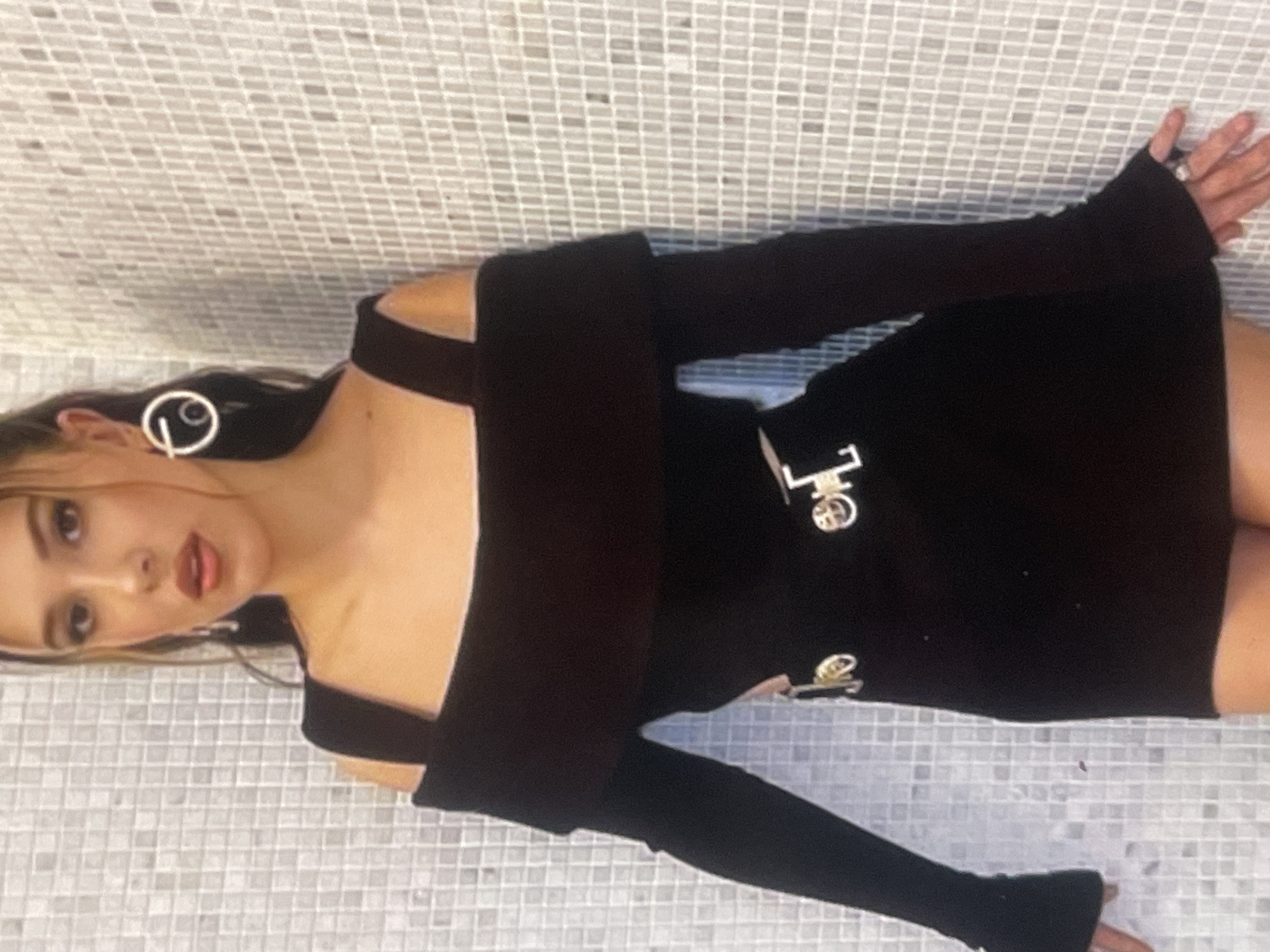 A young woman, likely in Poltava, Ukraine, poses for a photo against a light-colored tiled wall or floor. She is rotated 90 degrees counter-clockwise in the image, appearing to recline on her side. She has long, dark hair and is wearing a black off-the-shoulder mini-dress with long sleeves that cover her hands, featuring thumbholes or similar openings.
Her face is turned towards the camera, with a hint of a smile or pout. She wears large, circular white earrings, one of which prominently displays a white "P" or similar stylized letter within its design. On her right hand, a ring is visible.
The black dress is adorned with several metallic embellishments on the torso area. A prominent, stylized metallic letter, resembling a "P" or an "L" with an integrated circular element, is visible. Below it, there is a smaller, circular metallic emblem, possibly an interlocking logo or badge. Her hands are placed against the tiled surface, suggesting she is leaning or pushing off it. The background consists of small, light-colored square tiles, creating a grid pattern.