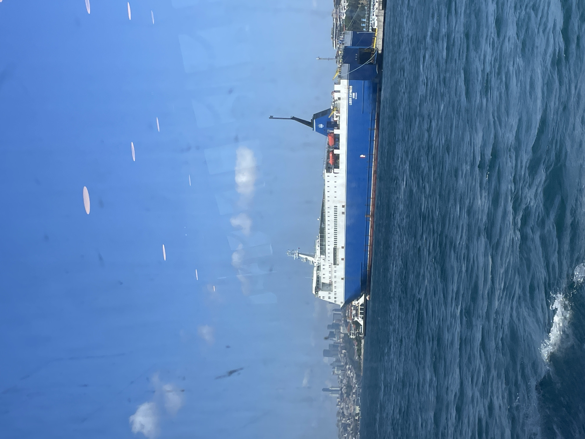 A wide-angle, outdoor, daytime photograph captures a large blue and white ferry docked at a pier in Üsküdar, Turkey. The ferry, named "LIDER EXPRESS," is positioned in the foreground, with its long hull extending across the frame. The water in front of the ferry is a deep blue with visible ripples. In the background, the Üsküdar skyline is visible, featuring numerous buildings and structures, including tall skyscrapers. The sky is bright blue with a few scattered white clouds. The photograph appears to be taken from another vessel or from a window overlooking the harbor. The lighting suggests it is a sunny day.