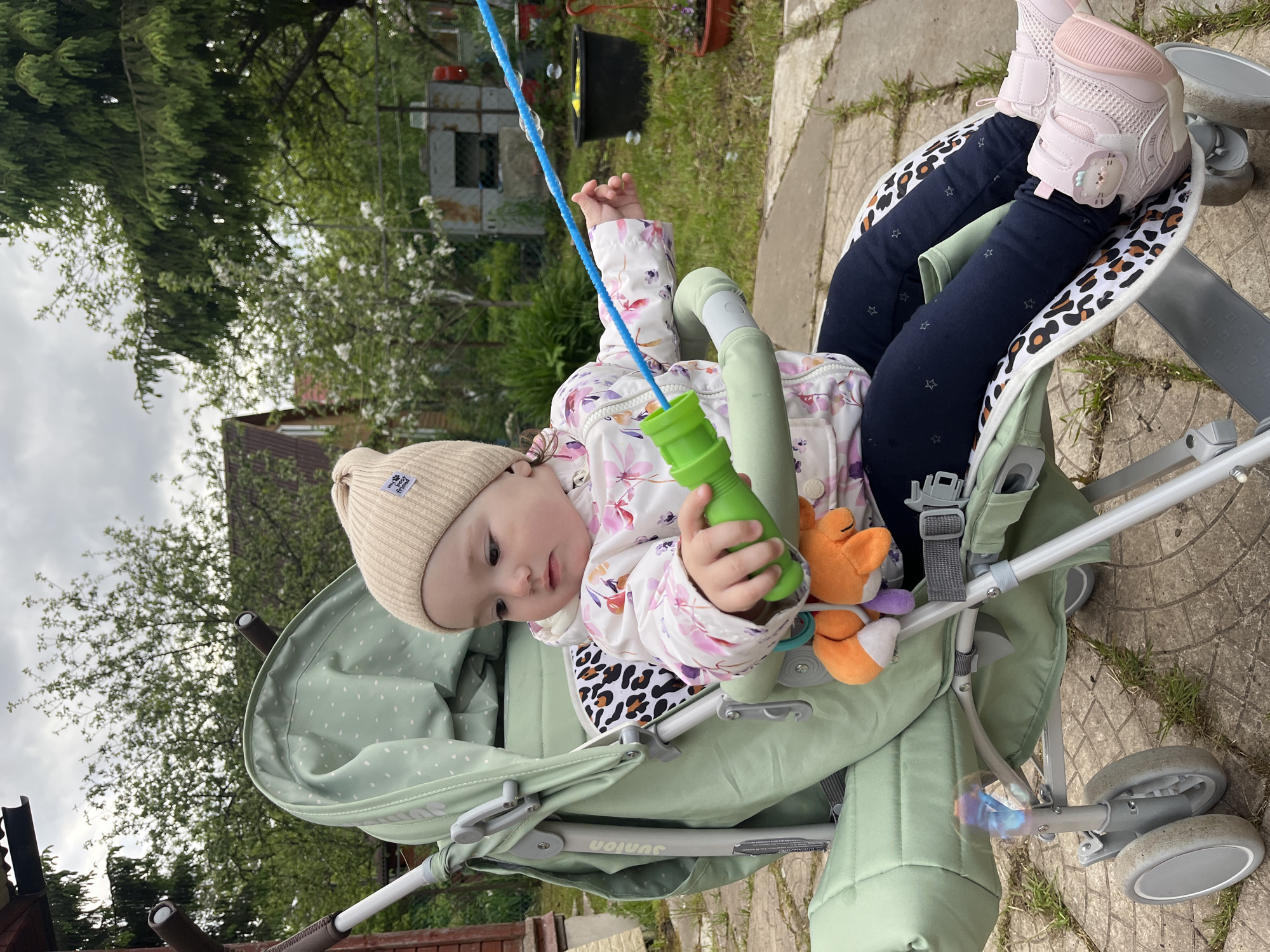 The image depicts a baby in a stroller in what appears to be a backyard or garden setting in Мшинская, Russia. The baby, wearing a beige knit hat and a floral patterned jacket over a leopard print inner layer, is holding a green bubble wand. The stroller is light green with a polka-dotted canopy. The baby's legs are clad in dark blue pants with star patterns, and pink sneakers. To the rear, the stroller's wheels are visible, as well as paving stones and patches of grass. Behind the stroller, there are trees, a fence, and parts of a house. A bubble wand is present near the baby. The sky appears overcast.
