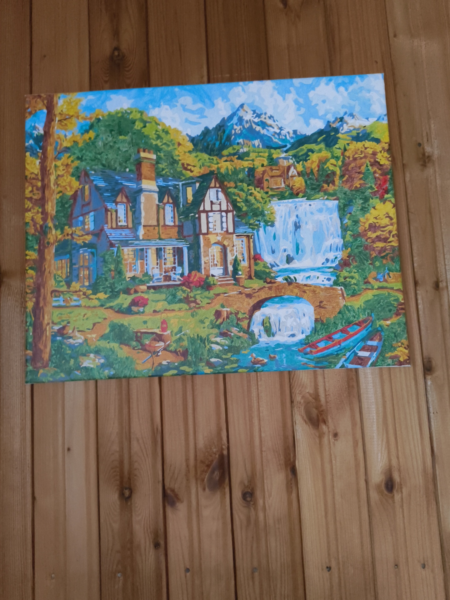The image is a painting depicting a picturesque landscape. In the foreground, a rustic scene unfolds with a stone bridge arching over a stream, leading towards a grand house nestled amidst lush greenery. The house, with its dark tiled roof and multiple chimneys, appears to be a substantial dwelling. The surrounding environment is vibrant with autumnal colors, indicated by the yellow and orange foliage of the trees. A cascading waterfall adds a dynamic element to the scene, with its white water tumbling into the stream below. Two canoes are visible in the water, one red and one white, adding a touch of human presence to the natural setting. In the background, majestic mountains rise, their peaks covered in snow, contrasting with the warm hues of the autumn landscape. The sky above is a brilliant blue, dotted with fluffy white clouds. The painting is displayed on a wooden wall, likely in a home, with the vertical planks of the wood visible. The painting itself is a paint-by-numbers artwork, evident from the distinct, blocky application of colors and the outlined sections characteristic of such kits. The overall impression is one of serene natural beauty and charming rural life. The location is stated as Востец Снт, Russia, however, without additional context or landmarks, it's impossible to pinpoint the exact geographical location within this area based solely on the painting.